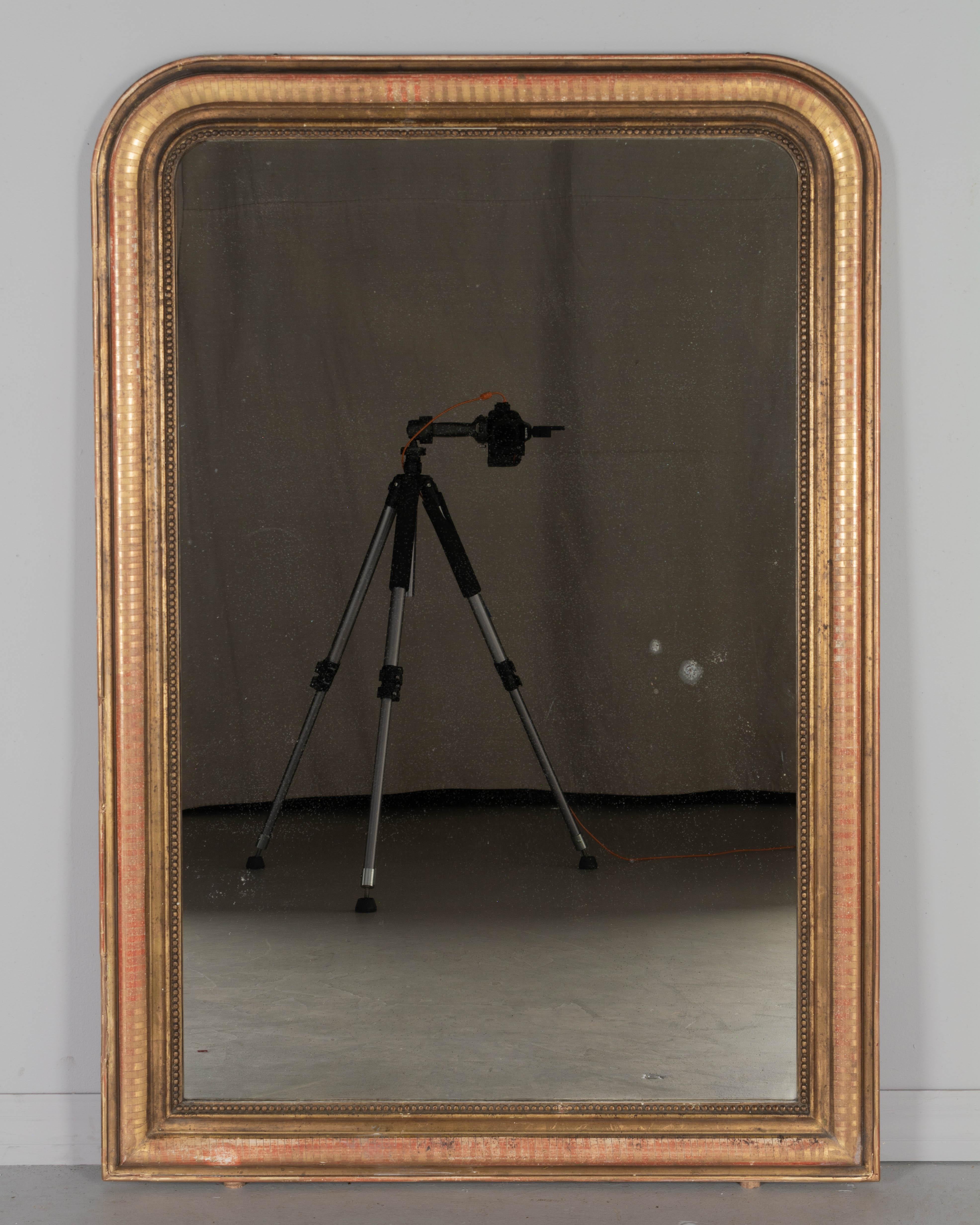 Miroir doré de style Louis Philippe, datant du début du 20e siècle, avec des angles supérieurs incurvés, un décor incisé et une bordure intérieure en perles. Finition dorée chaleureuse avec exposition du rouge sous le frottement. Miroir original