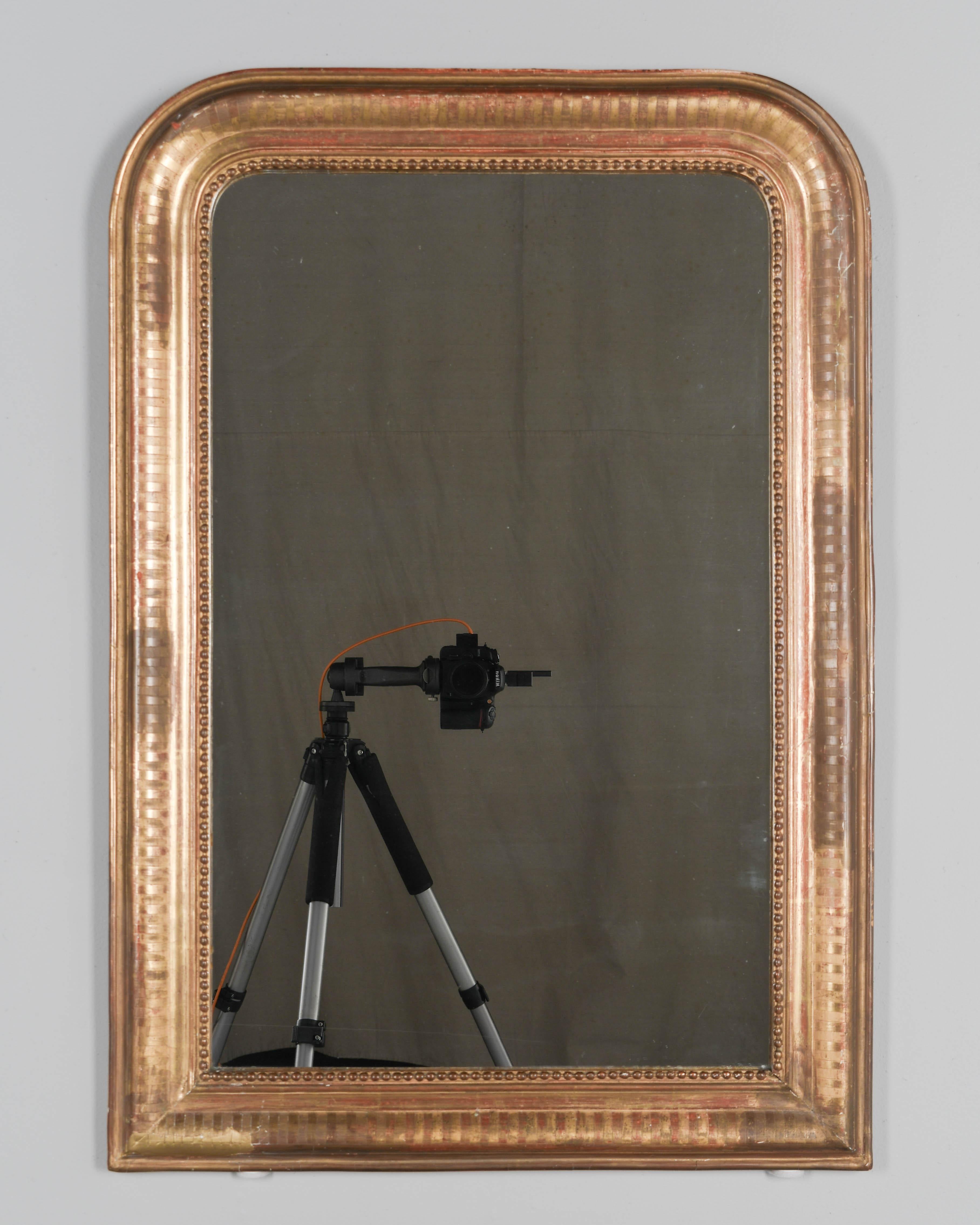 An early 20th century French Louis Philippe style gilded mirror with curved top corners, incised decoration and inner bead border. Warm gilt finish with red under rub exposed. Original mirror with some spotting. Frame has been restored, mainly on