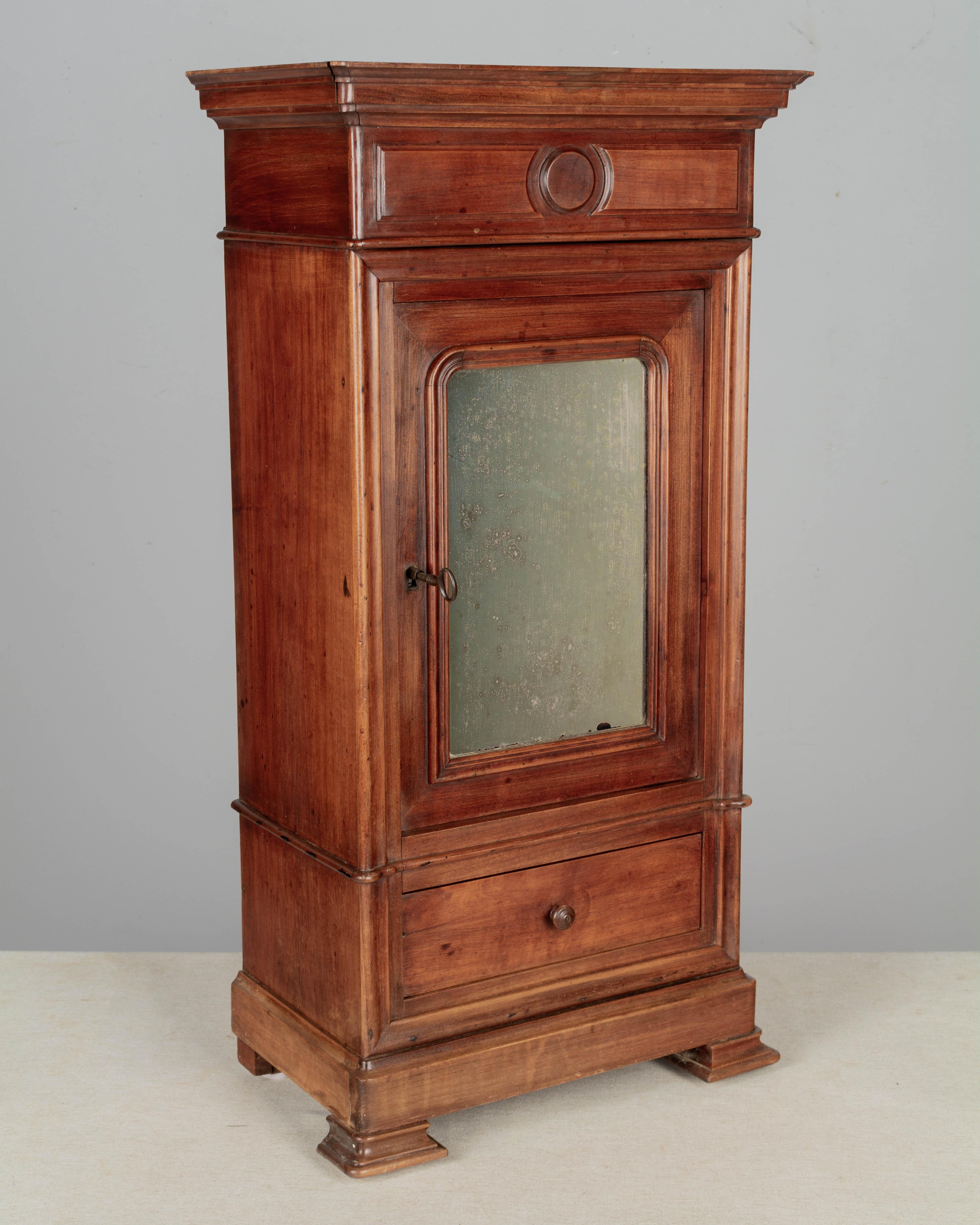 A 19th century Century French Louis Philippe style miniature armoire or sample cabinet, hand-crafted of solid walnut with a mirrored door and dovetailed drawer. Interior has one adjustable shelf (replaced). Working lock and key. Chestnut as a