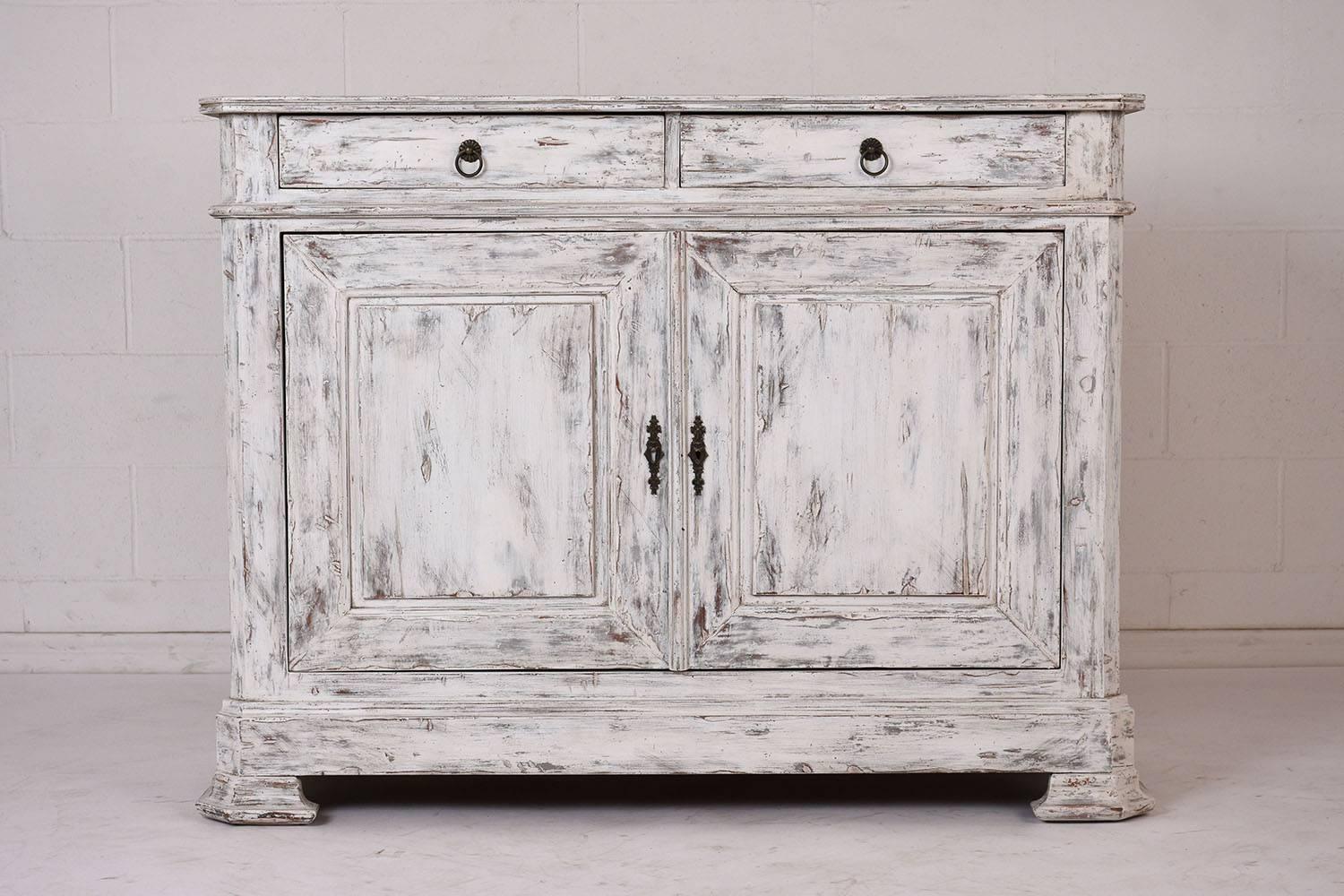 This 1970s French Louis Philippe-style buffet is made of mahogany wood painted in a off white and grey color combination with a distressed finish. There are two drawers at the top with decorative brass pulls. Below there are two cabinet doors with a
