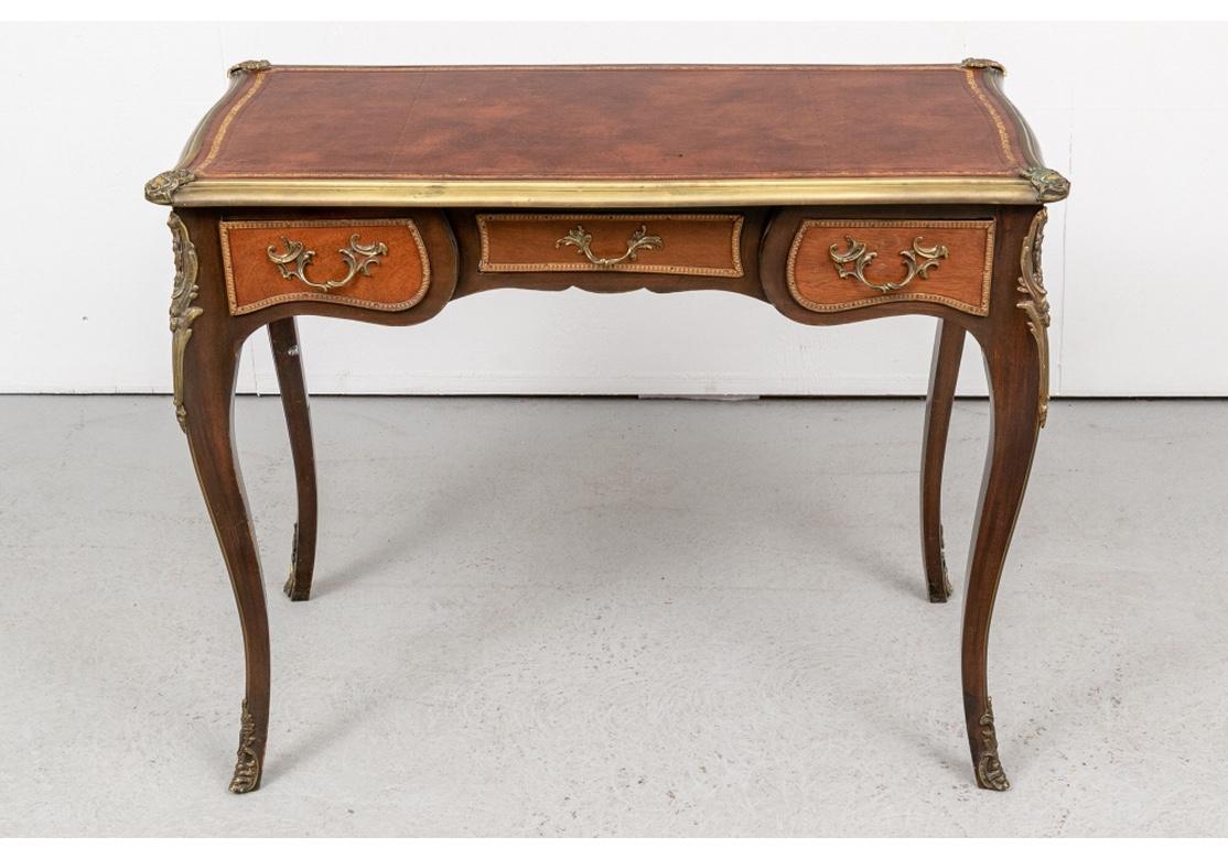 Made in France. A Louis XV style desk with brown tooled leather shaped top and brass banded edge with shell form mounts on the corners. The dark wood frame with lighter wood for the three front apron drawer panels with their brass egg-and-dart trim.