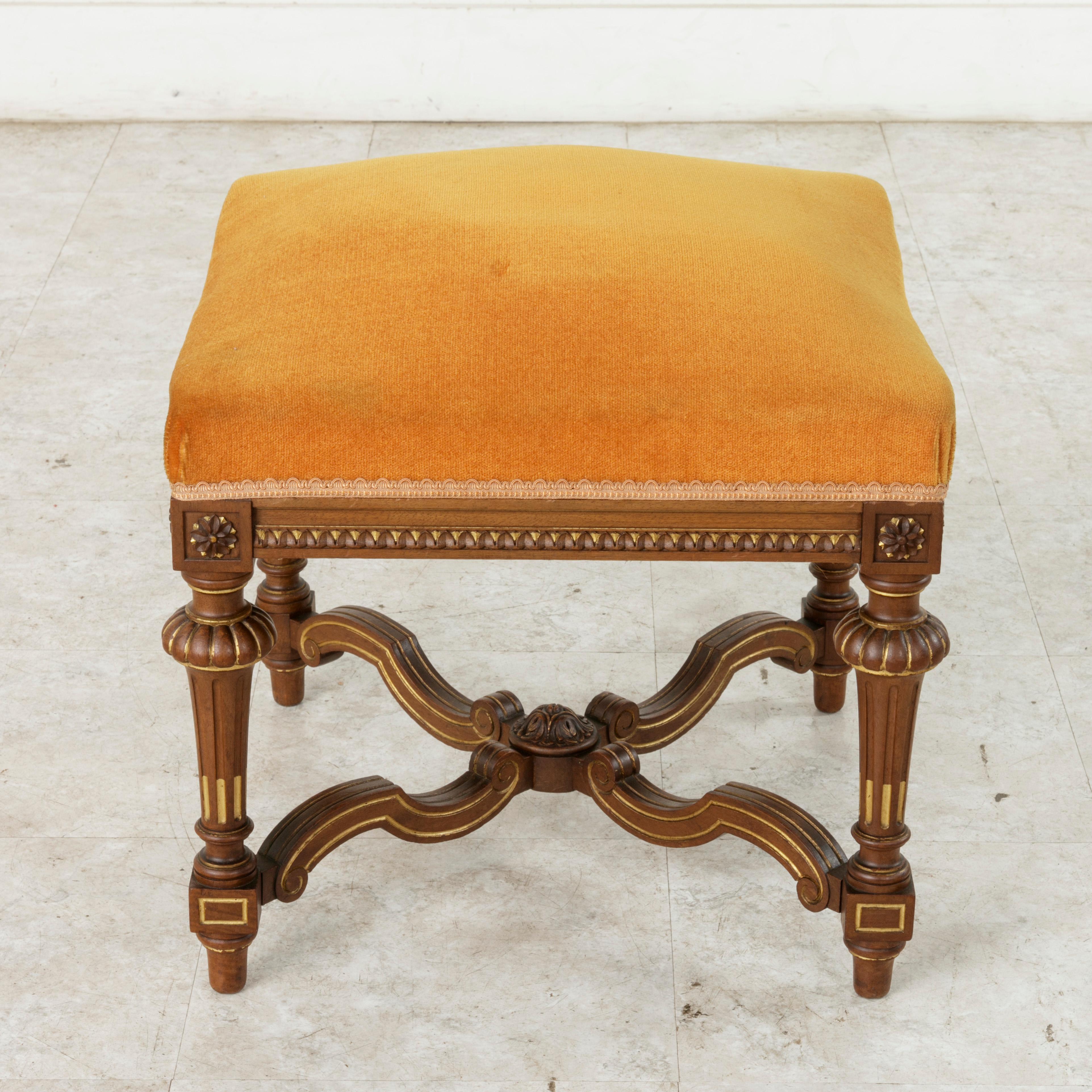 This turn of the 20th century French Louis XIV style walnut banquette, stool, or ottoman features hand painted gold detailing on the legs and stretchers. Carved details of rosettes adorn the die joints and carved rais de coeur surrounds the lower