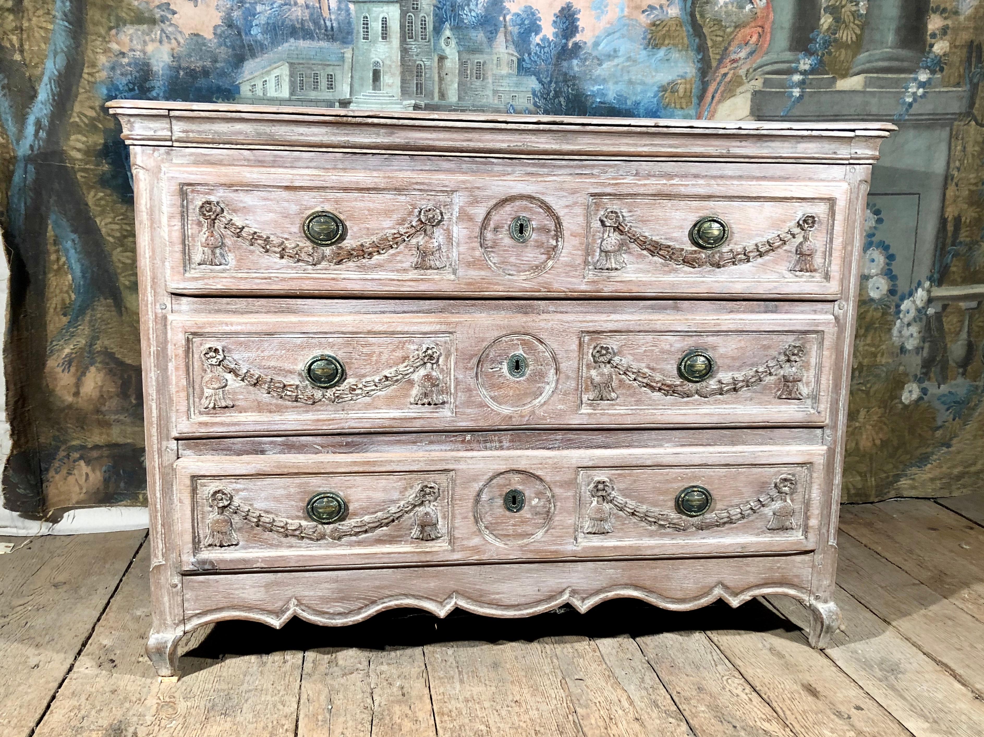 A late 18th century three-drawer commode in bleached oak, with its original wood top and brass drawer pulls, transitional Louis XV-Louis XVI, with wonderful garland carvings on drawer fronts, circa 1780.