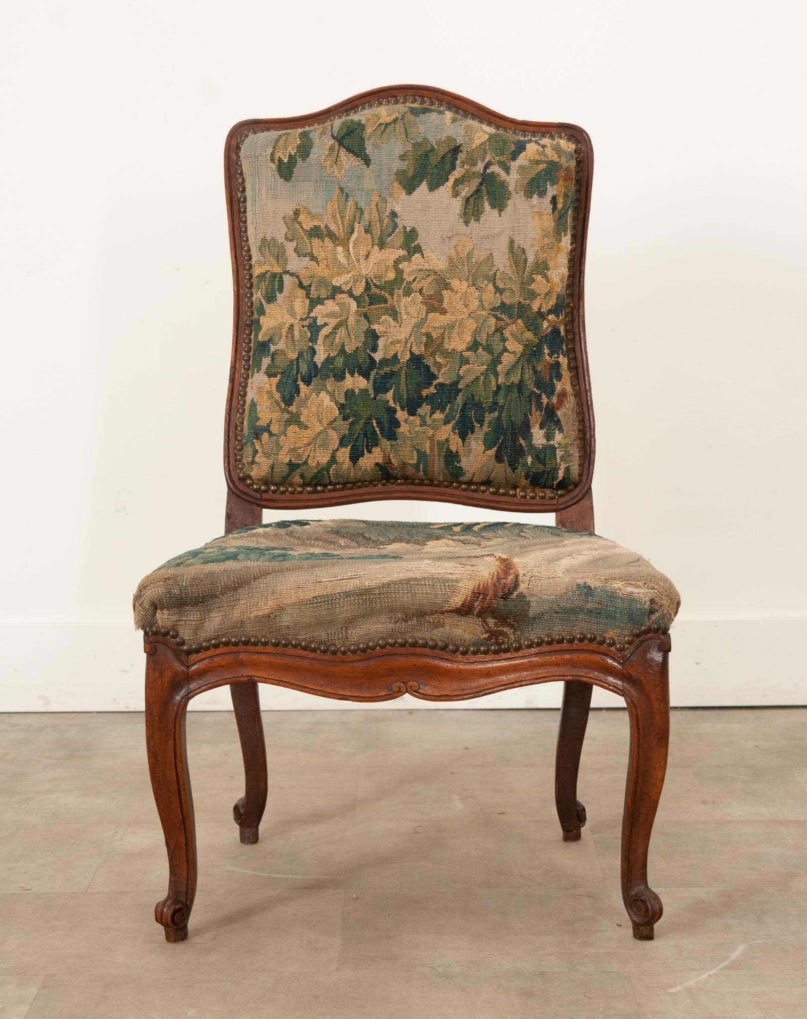 A charming and quite comfortable French 18th Century Louis XV period side chair with original tapestry. A camel hump top rail follows down to a shaped seat above a channel carved walnut scalloped apron ending in cabriole legs and scrolled feet.