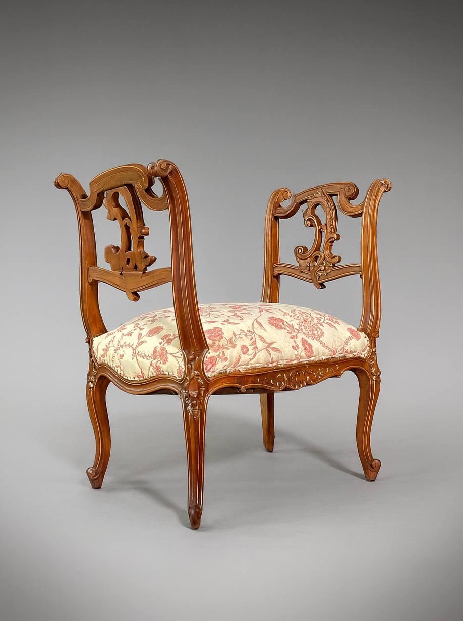 Conditio sine qua non, tappezzeria nuova raccomandata 

Questa panca da toeletta da boudoir in stile French Rococo XV presenta una splendida struttura in noce intagliata con bracci a spirale, motivi floreali e sottili accenti dorati che