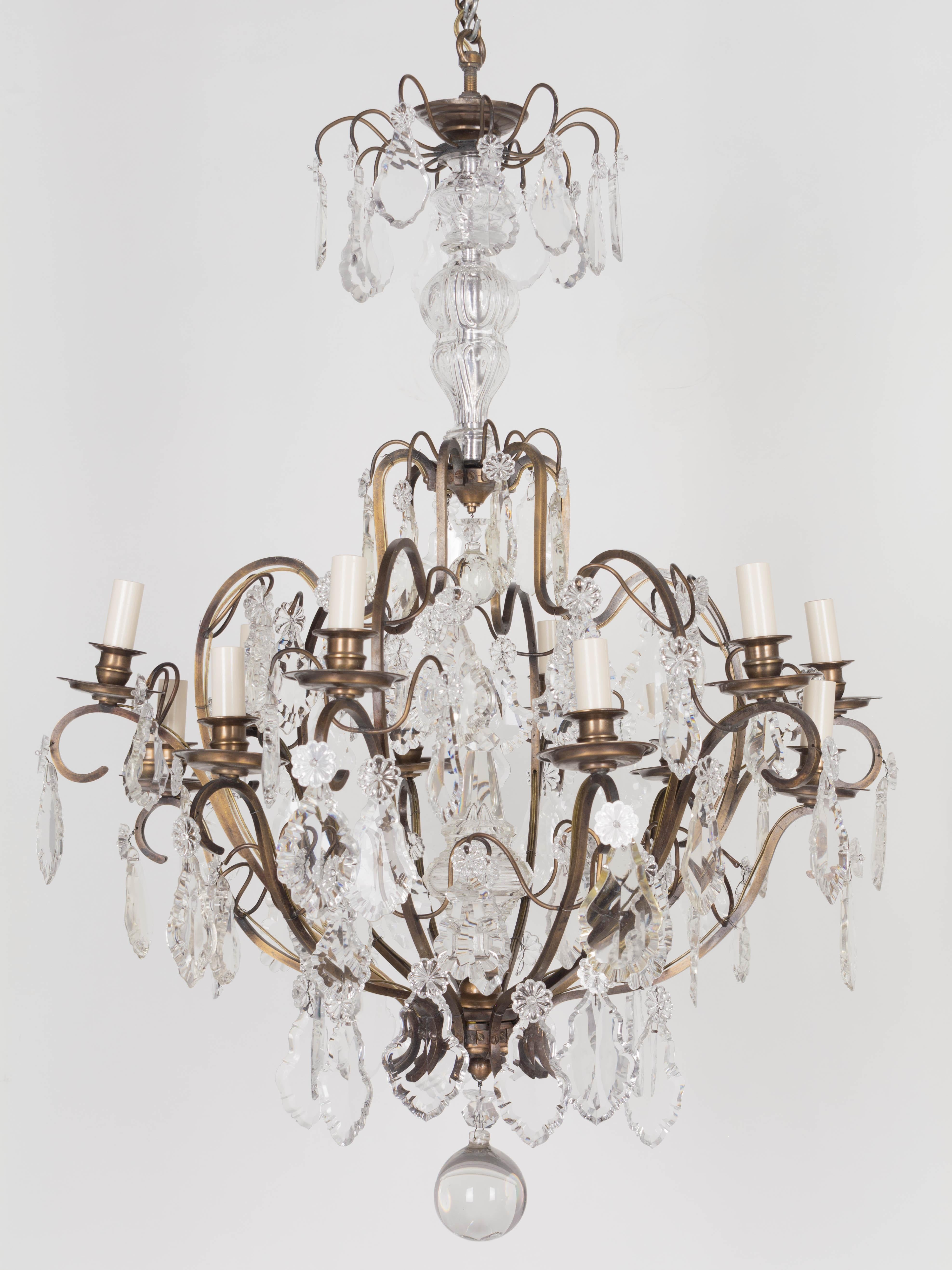 Lampadario a 12 luci in cristallo in stile Luigi XV dell'inizio del XX secolo con una serie di pendoli in cristallo con rosette. Telaio in ottone massiccio con bracci a voluta, bobeches in ottone ed elementi decorativi. Grande picco di cristallo al