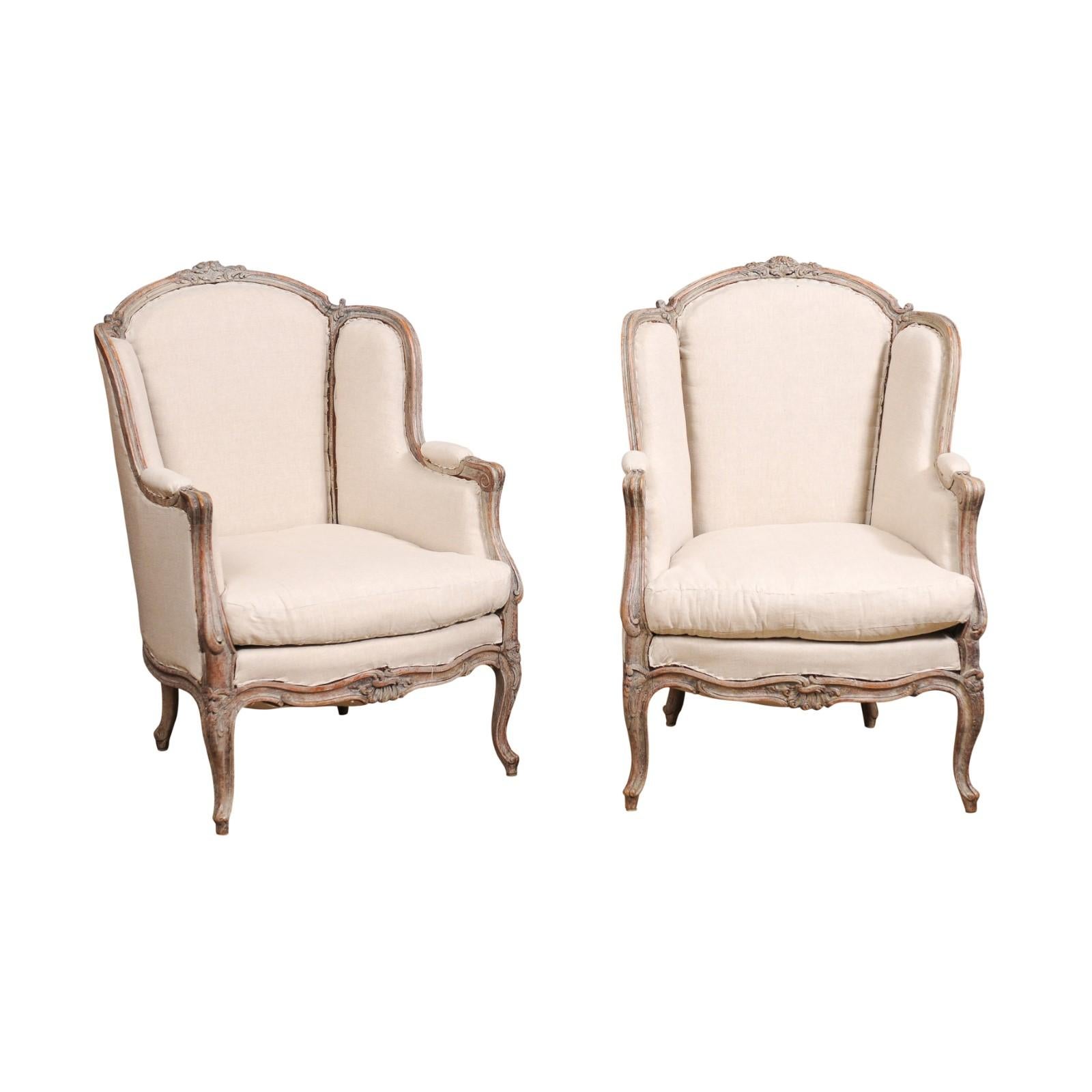 A pair of French Louis XV style painted wood bergères à oreilles chairs from circa 1880 with carved floral motifs, cabriole legs and used upholstery. This exquisite pair of French Louis XV style bergères à oreilles, dating back to circa 1880,
