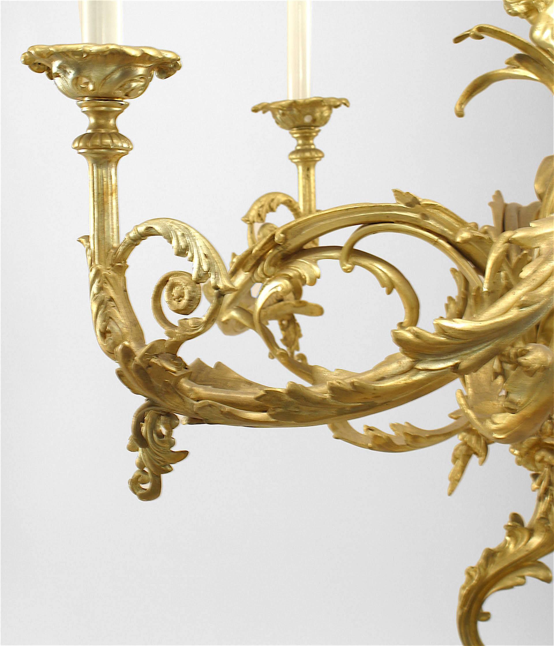 Französischer Kronleuchter aus vergoldeter Bronze im Louis XV-Stil (19./20. Jh.), mit 4 Rollenarmen und 2 Amor-Figuren auf einem zentralen Schaft mit Rollen oben und unten.
