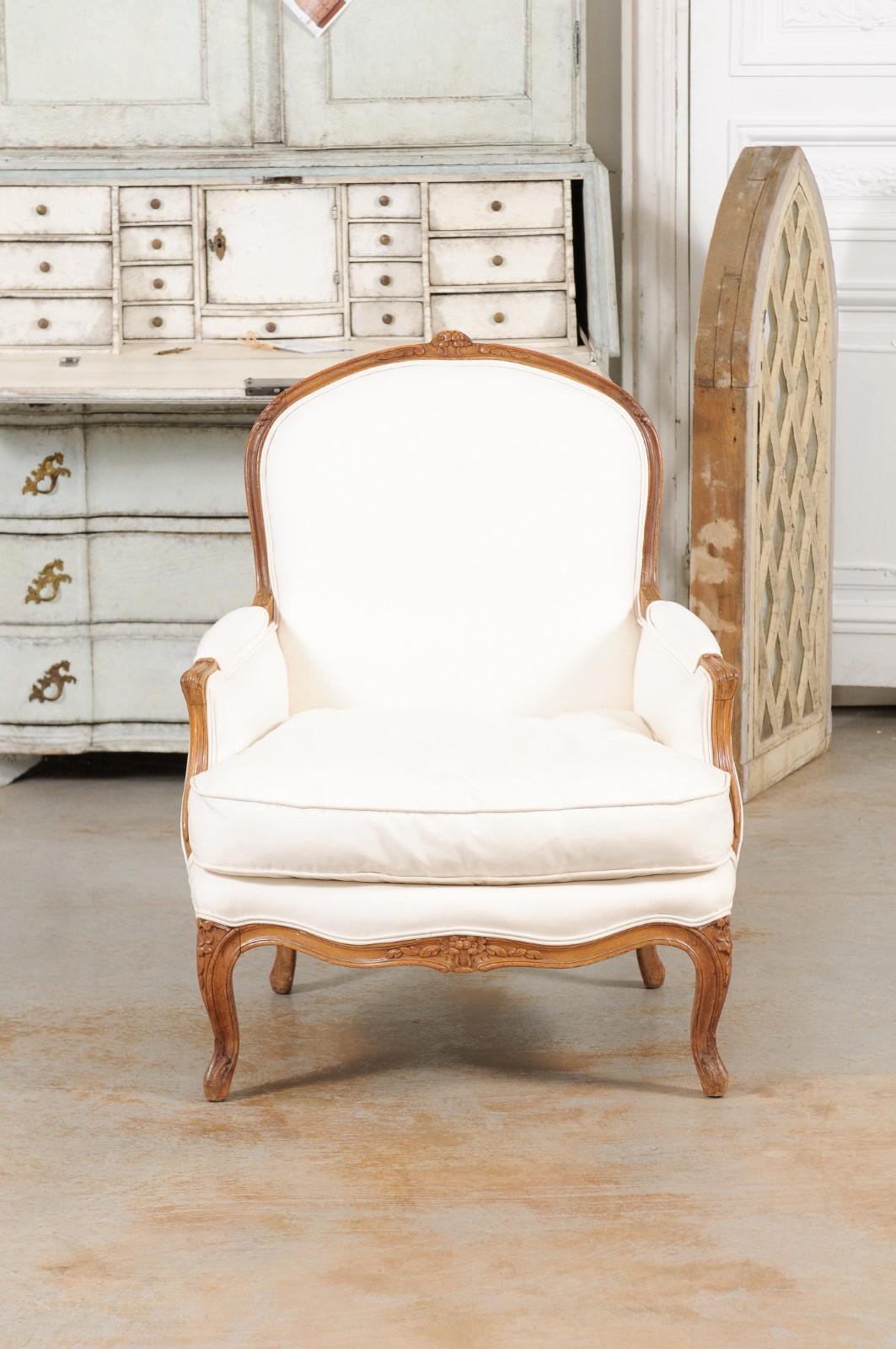 A French Louis XV style bergère chair from the 19th century, with carved floral motifs and new upholstery. Created in France during the 19th century, this Louis XV style wooden bergère features a slightly slanted back with carved flowers on the