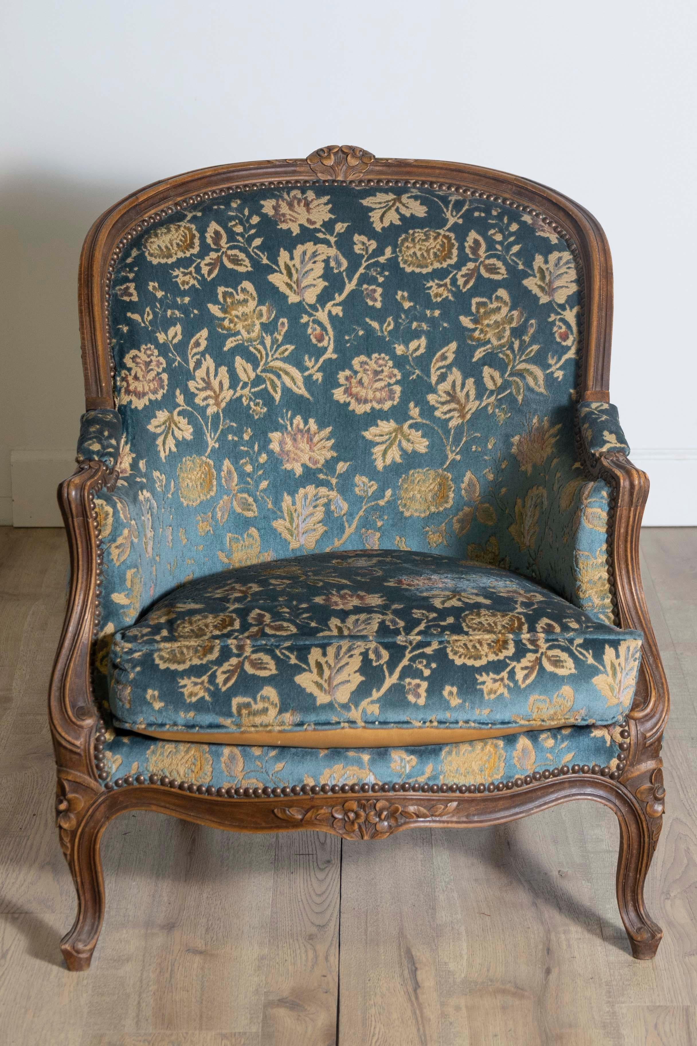 A French Louis XV style walnut bergères chair from the 19th century with carved crests, scrolling arms, floral carved apron, cabriole legs and floral upholstery. Enhance your living space with this exquisite French Louis XV style walnut bergères