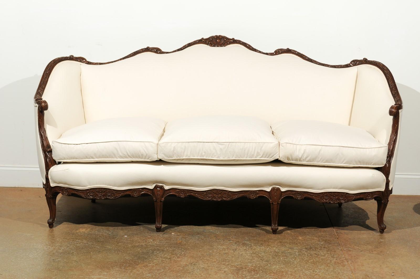 A French Louis XV style walnut three-seat sofa from the 19th century, with hand-carved crest and skirt, floral décor, scrolling arms, cabriole legs and new upholstery. This French sofa features a beautifully serpentine upper rail, adorned with