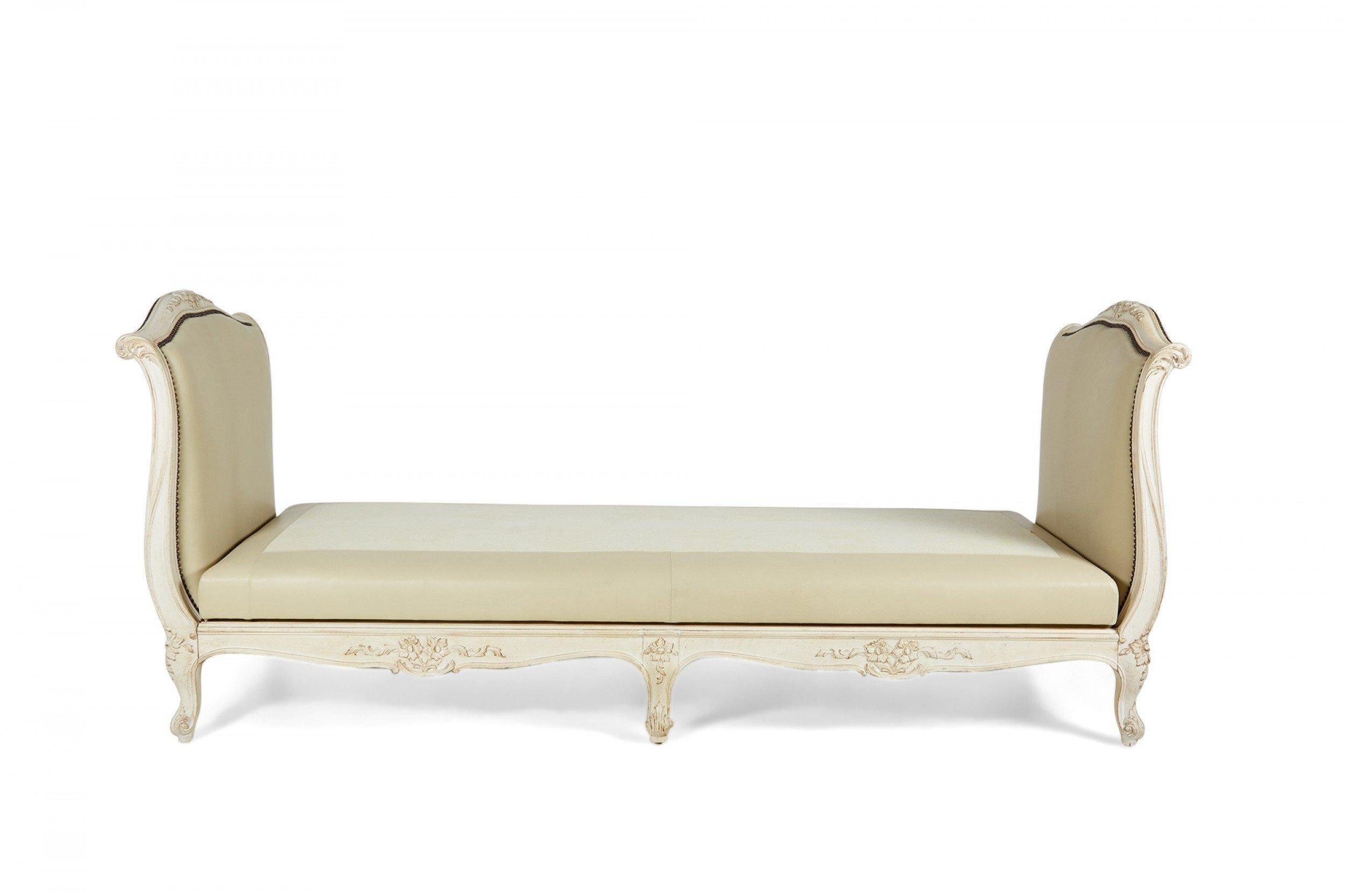 French Louis XV-style (1st half 20th Century) daybed with white distressed painted finish having carved embellishments, and beige leather upholstered headboard, footboard, and base adorned with decorative furniture tacks. (Does not include seat