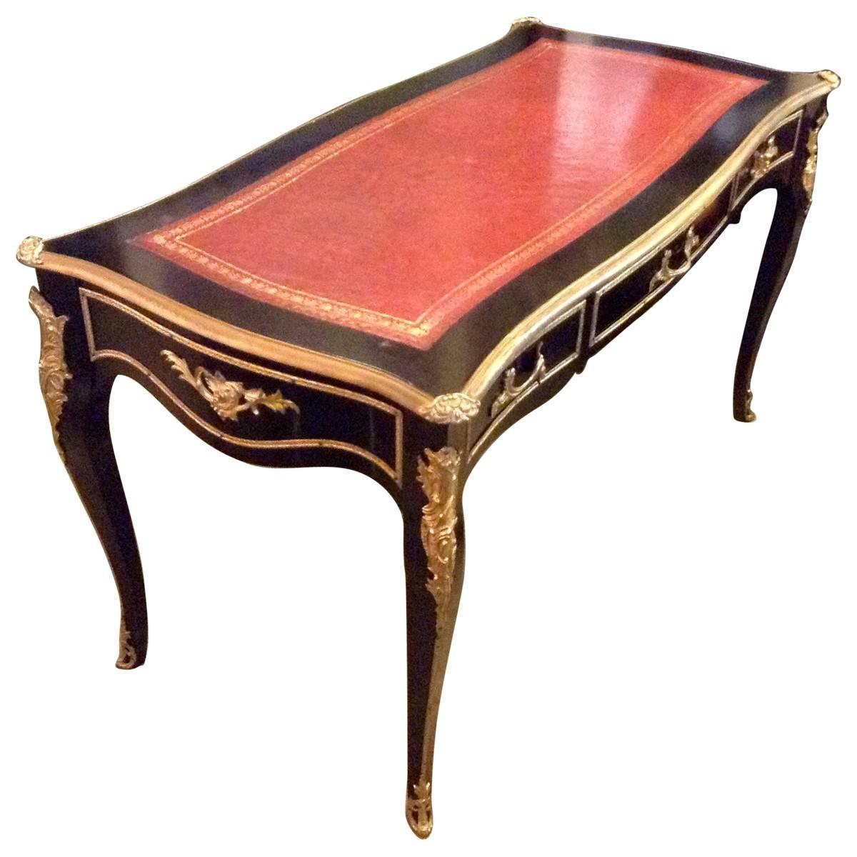 This antique bureau plat was designed and handcrafted in France in the luxe style of Louis XV. Its black lacquered hardwood body is accented with delicate gilt detail with a top inlaid with sumptuous red stamped leather. Cabriole legs and three