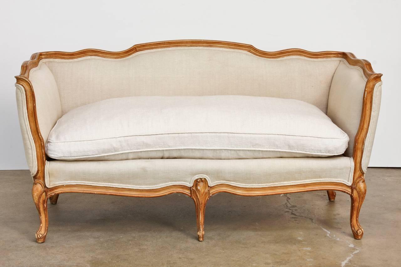 Classic French Louis XV style canape settee featuring a hand-carved frame. The settee is upholstered in a thick linen with a later loose cushion in a similar shade and texture. The linen fabric is bordered with a double welt and is showcased by the