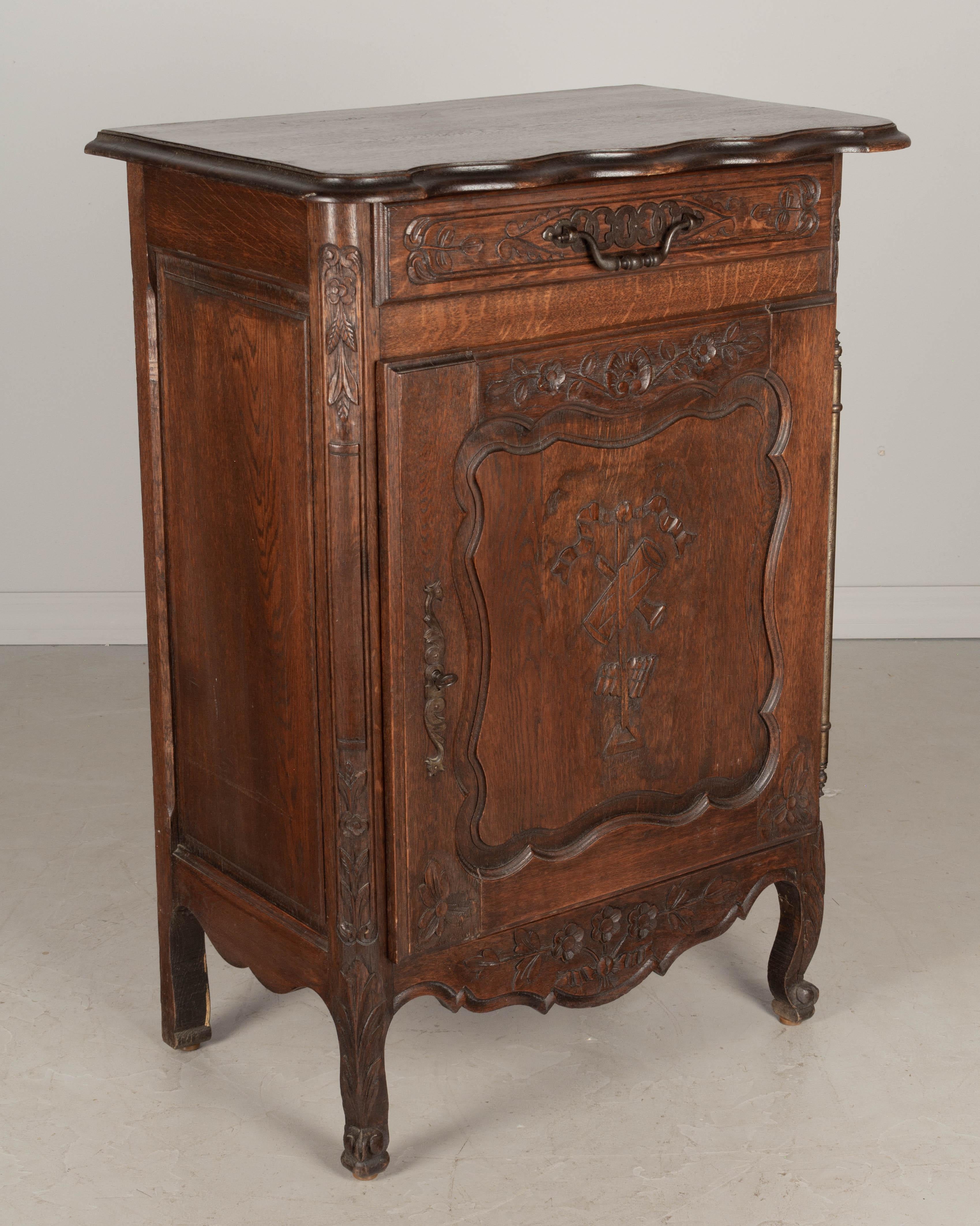 A Country French Louis XV style confiturier or single door cabinet from Normandy. Made of solid oak with hand-carved floral details on the corners and apron and musical instruments on the door. Dovetailed drawer with iron pull. Cabinet door has