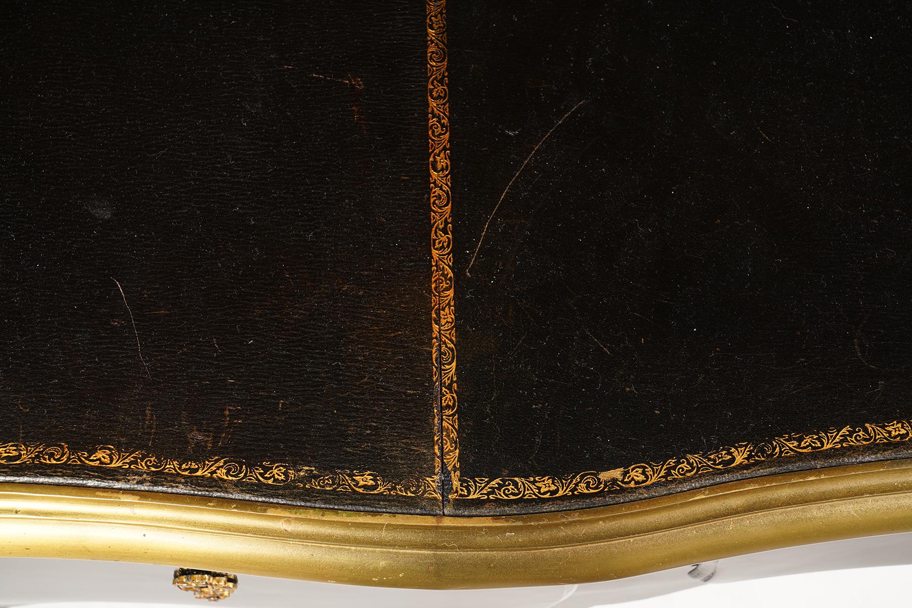 This elegant vintage French ebonized and ormolu-mounted bureau plat, dating to the late 20th century, features a gilt tooled black leather writing surface surrounded by a gilt bronze band with accented corners above a serpentine shaped frieze with