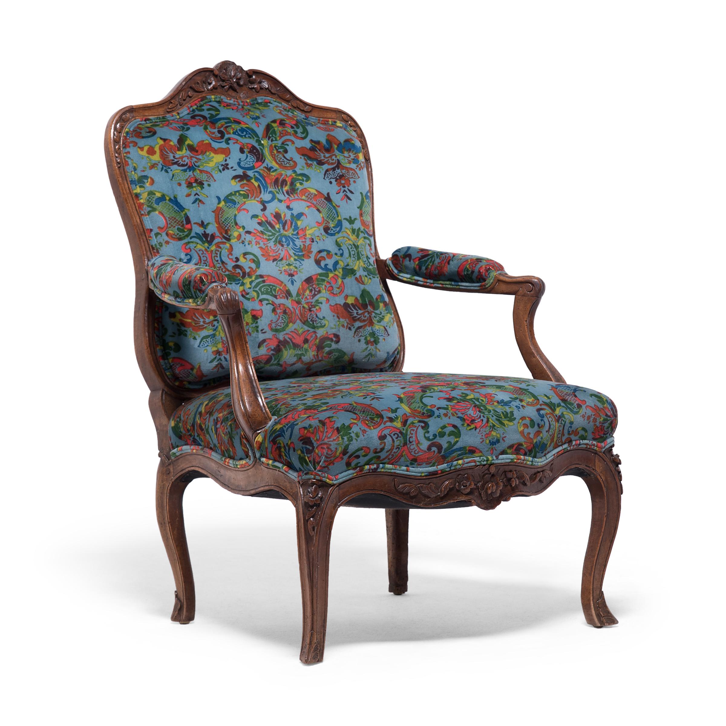 This 19th-century French upholstered armchair wonderfully embodies the Louis XV style with rich ornamentation and nary a straight line in sight. The chair has a sinuous wooden frame with a wide seat, flat back, and open, padded armrests. The crest