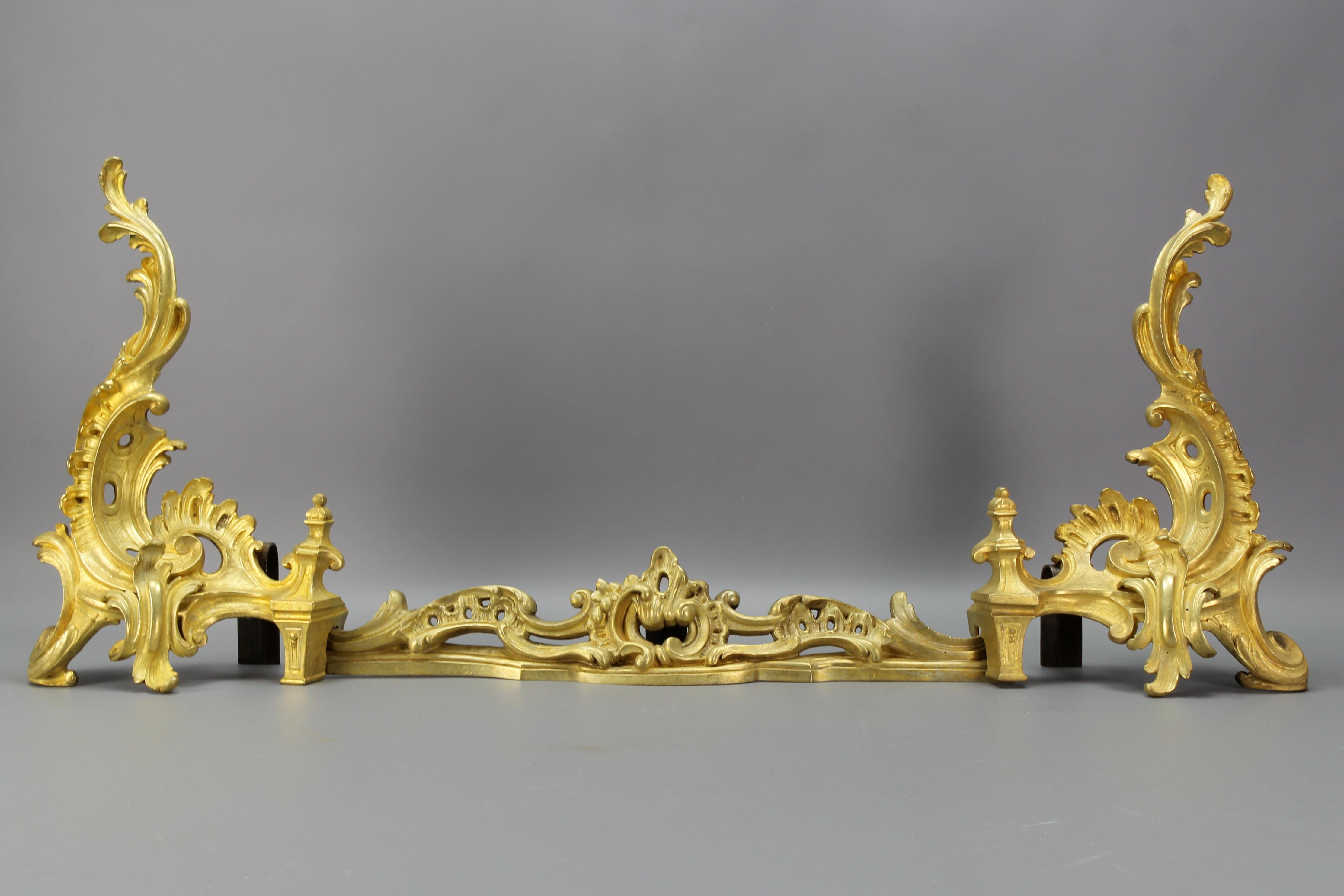 These impressive French Louis XV or Rococo-style gilt bronze andirons with their crossbar date to the end of the 1800s. 
Richly ornate with rocailles, C-scrolls, and foliate accents, with very curved edges. 
This beautiful antique fireplace set has