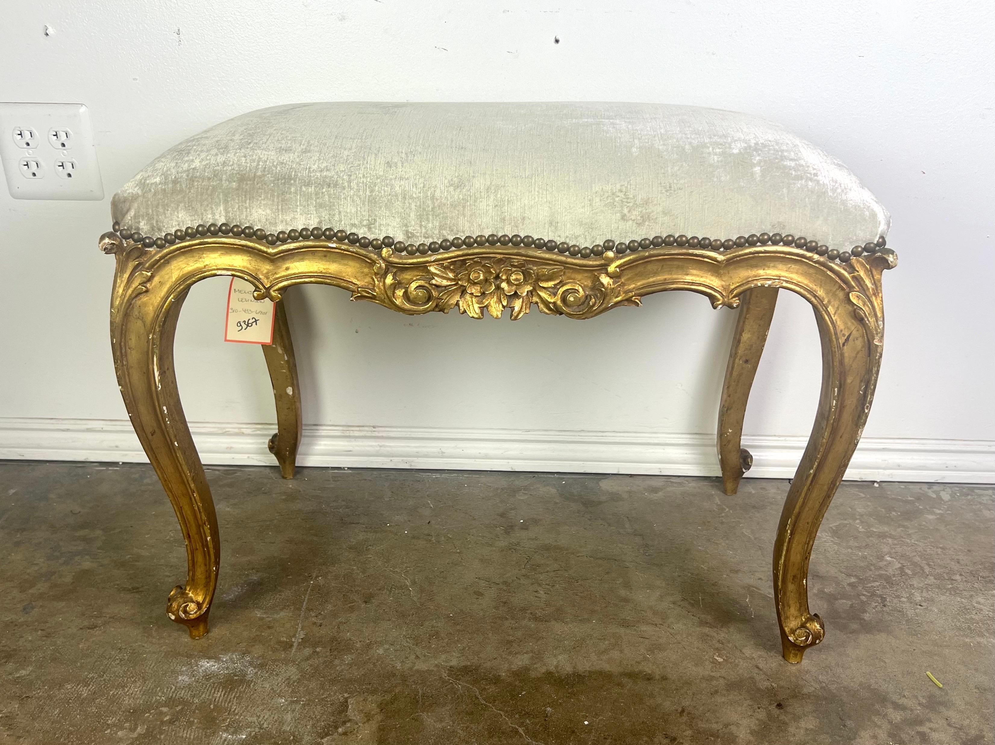 This bench is an elegant piece featuring a French inspired design with opulent details.  The frame is crafted from wood with a gold gilt finish that accentuates its ornate carvings and floral motifs, particularly visible along the apron and the