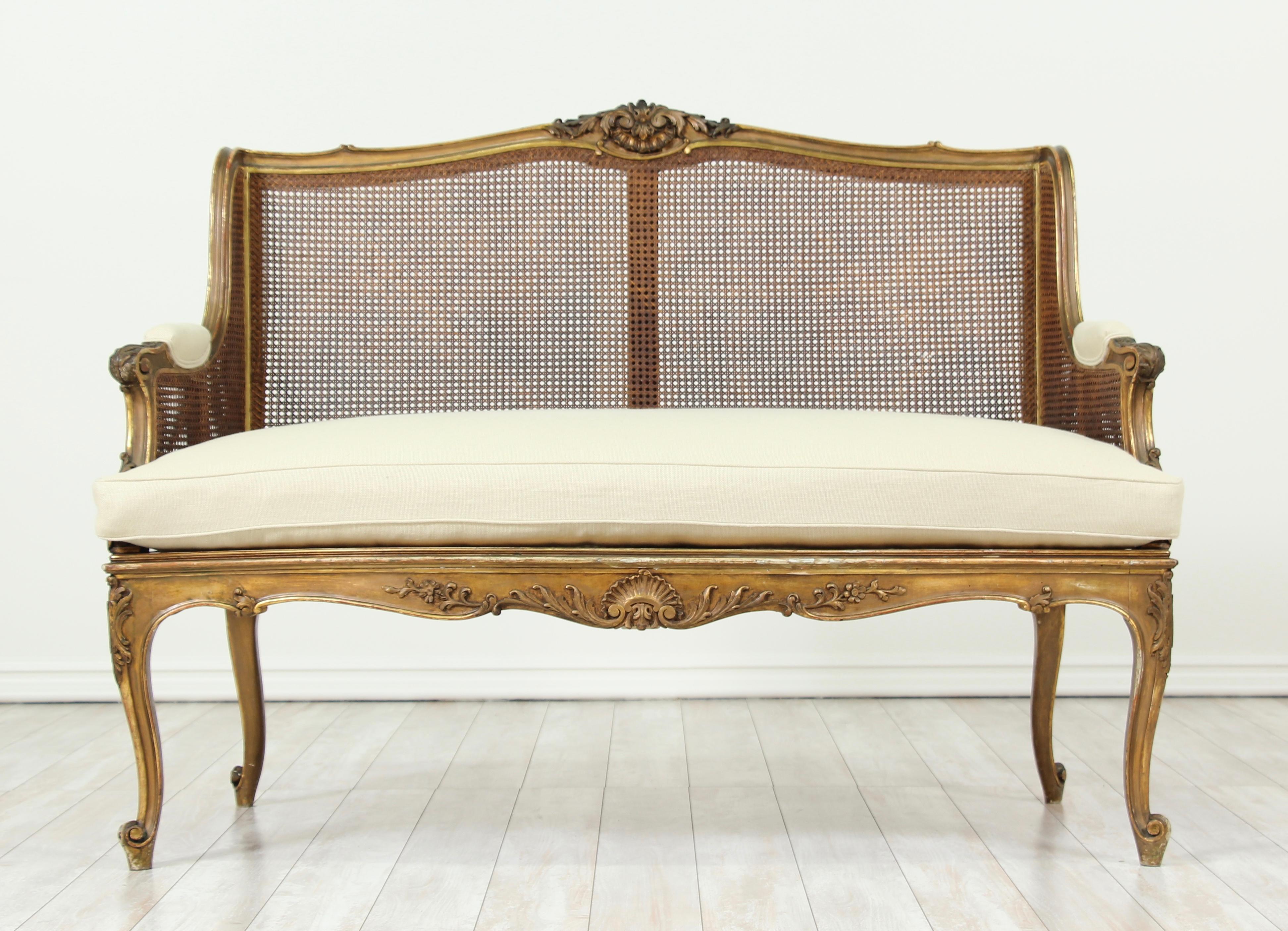      Beautiful, 1920s French Louis XV-style caned giltwood settee with delicately carved decorations and new Belgian linen upholstery (loose seat cushion). 
     The settee would add a wonderful sense of French airiness to any interior. 
    