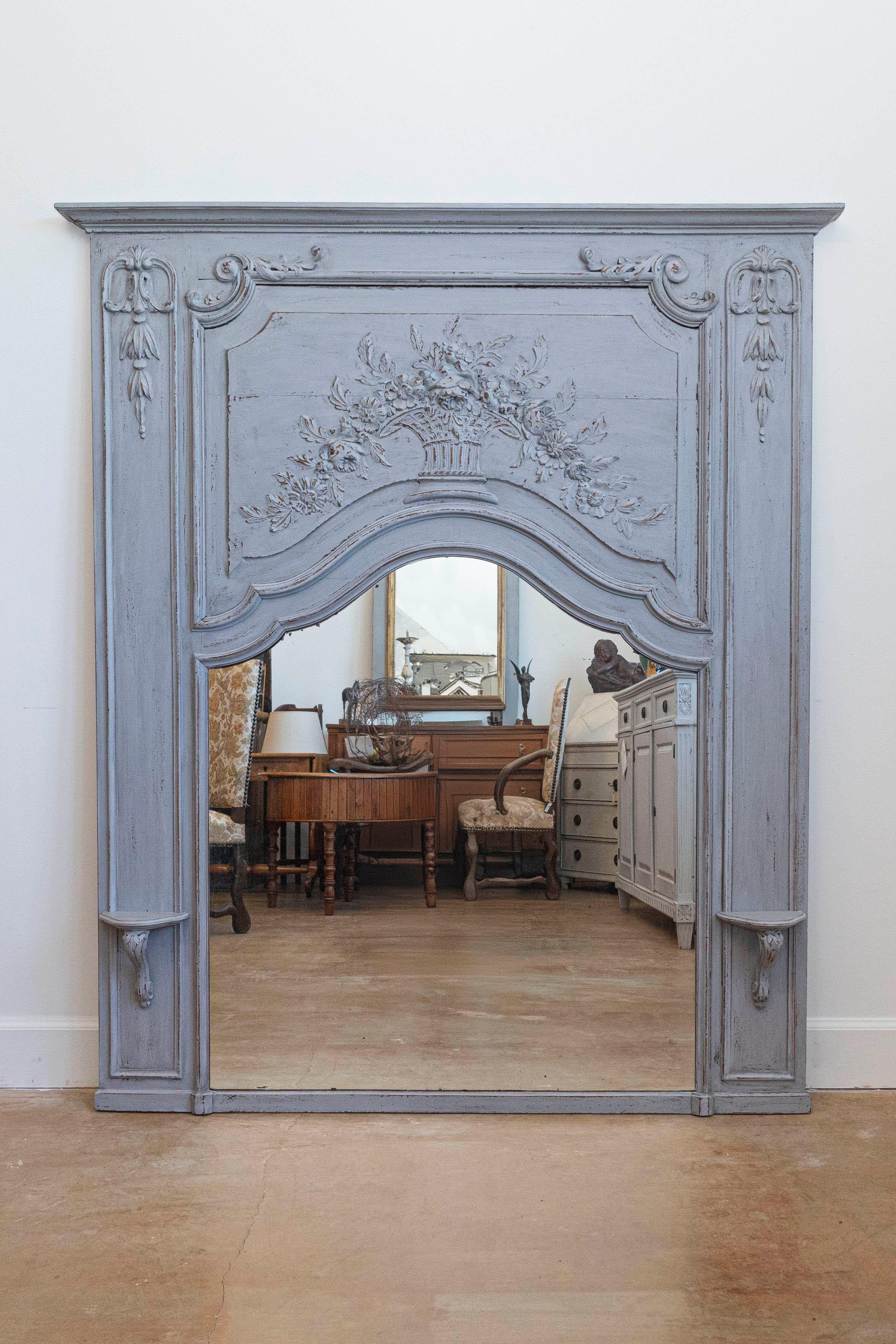 A French Louis XV style grey painted wood trumeau mirror from the 20th century, with carved bouquet in wicker basket and brackets. Born in France during the 20th century, this exquisite trumeau mirror showcases the stylistic characteristics of the