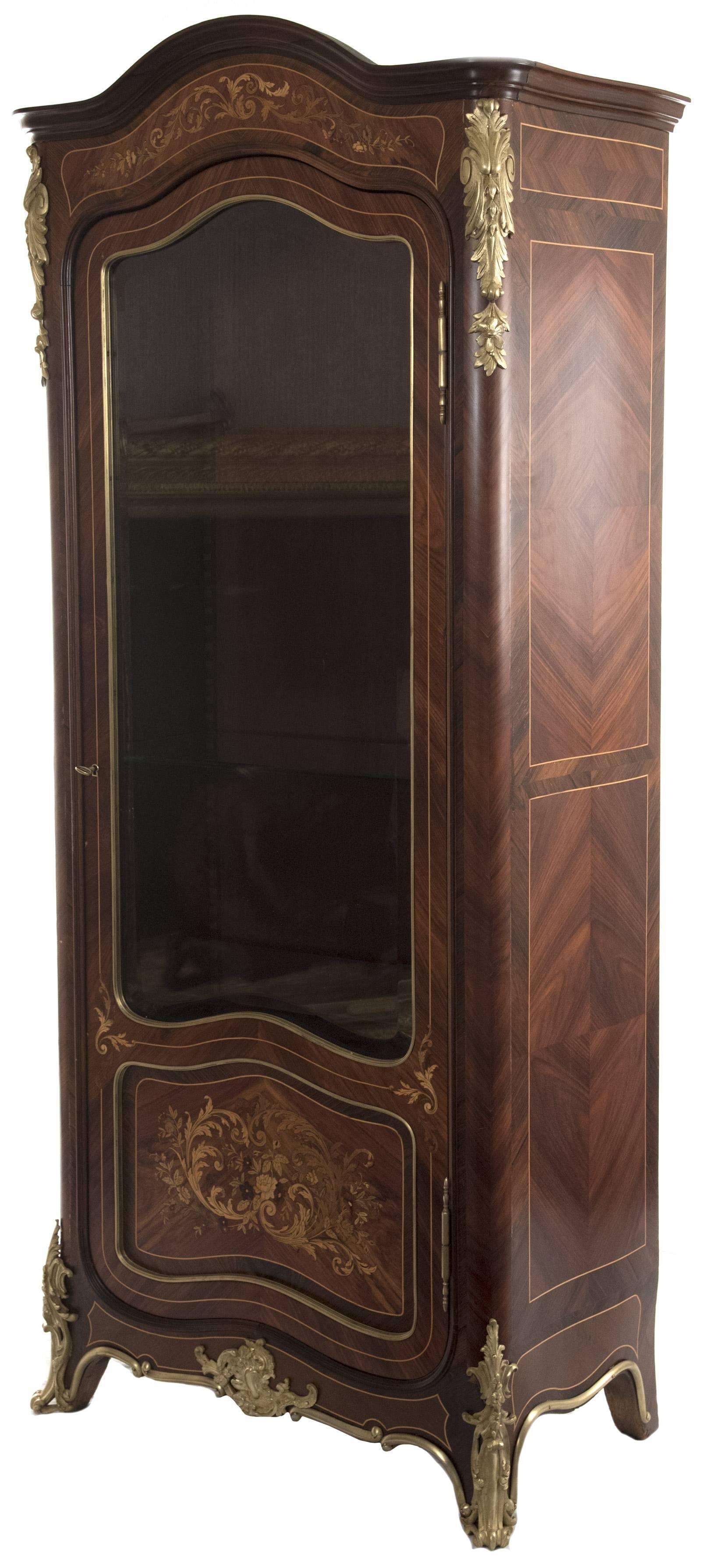 A French Louis XV style vitrine (Napoleon III) covered in figured veneer panels with inlaid details in contrasting woods. Below the rounded pediment is an inlaid panel of conforming shape with a delicately scrolling foliate motif, above a glazed