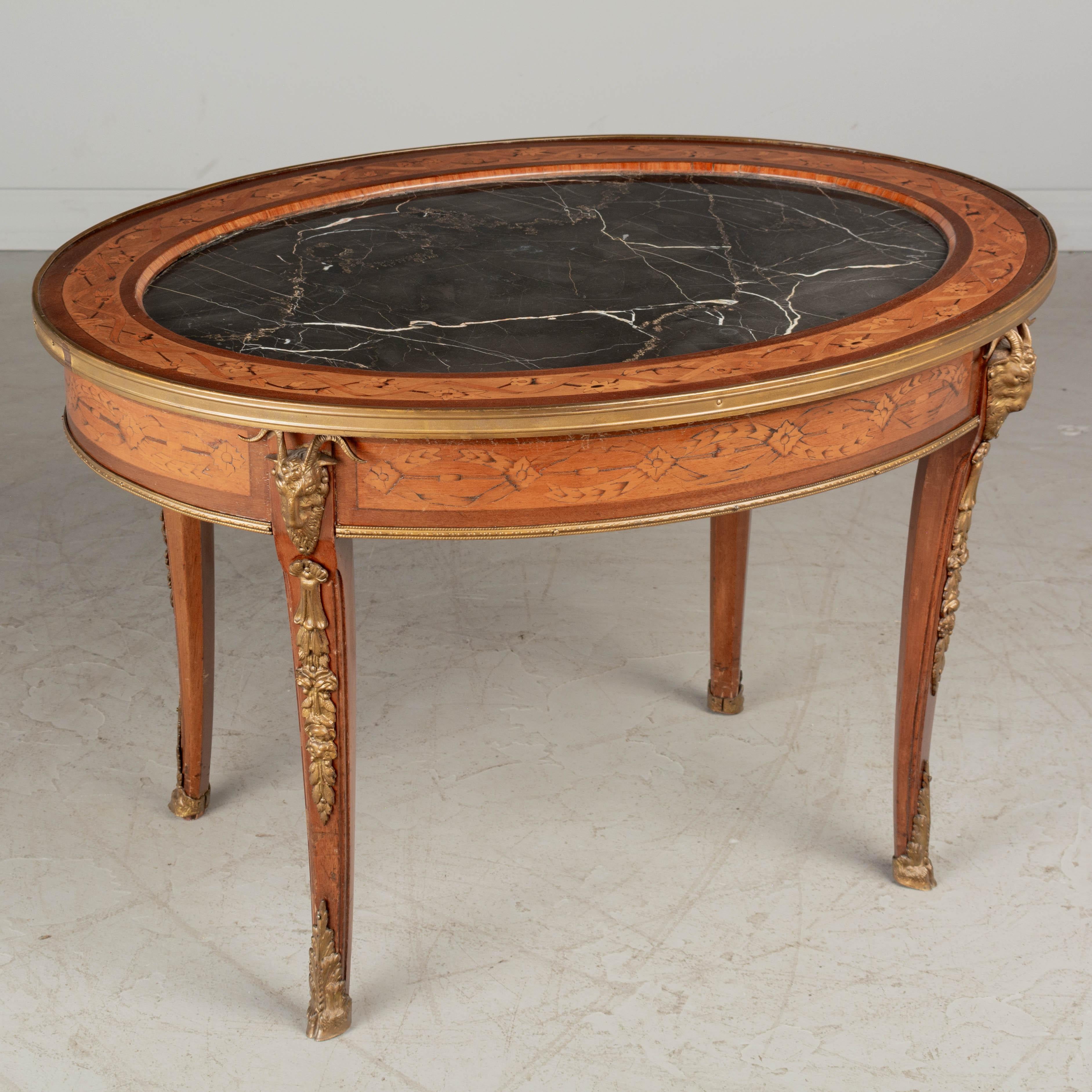 A French Louis XV style oval coffee table with marquetry veneer of mahogany and inset black marble top. Cast bronze decorative details and brass trim. All original. Circa 1920s. 
Dimensions: 30.25