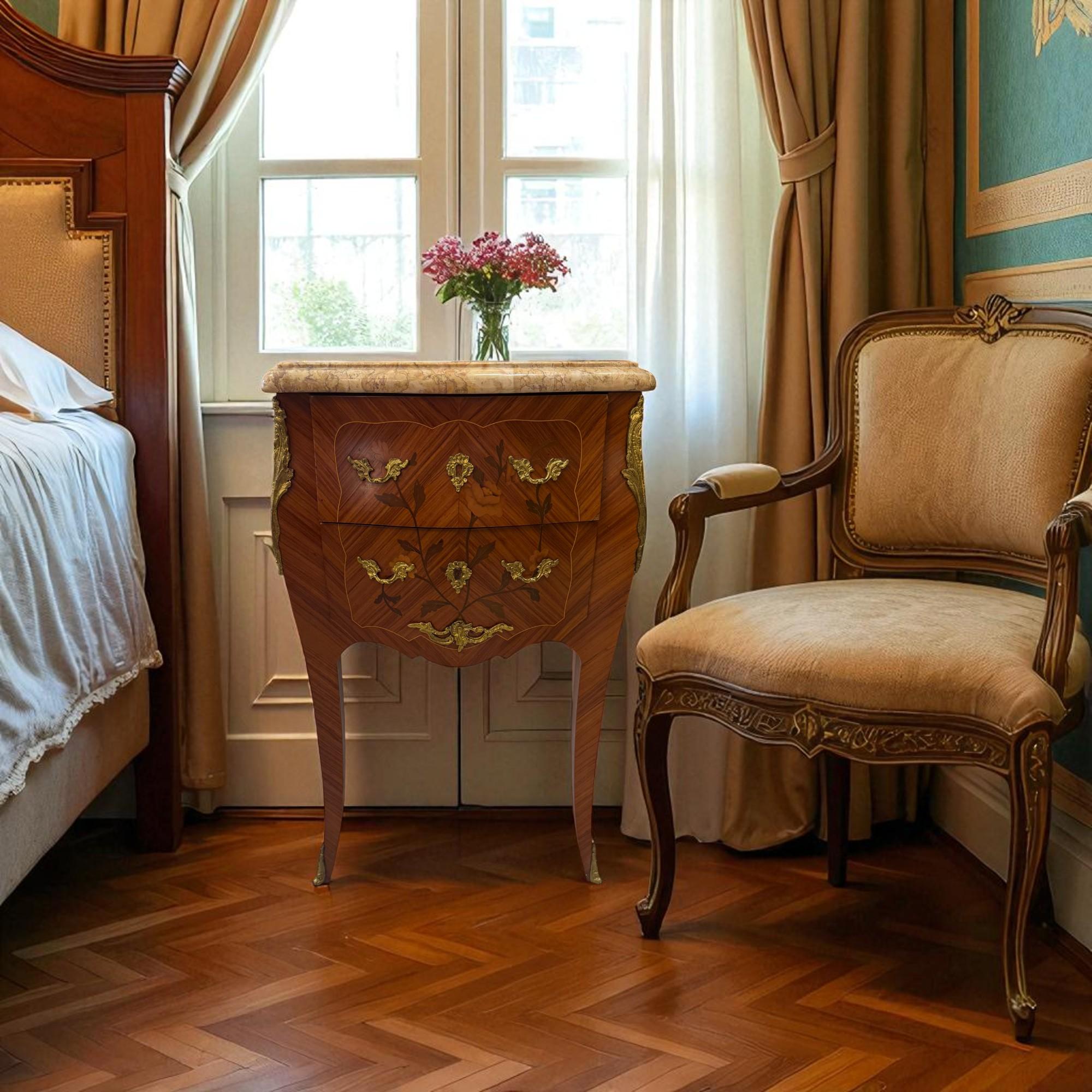 This two-drawer bedside table or end table is a fine example of French craftsmanship in the Louis XV style. The piece features a beautifully shaped body with delicate floral marquetry inlays set against a warm wood veneer. Intricate bronze mounts