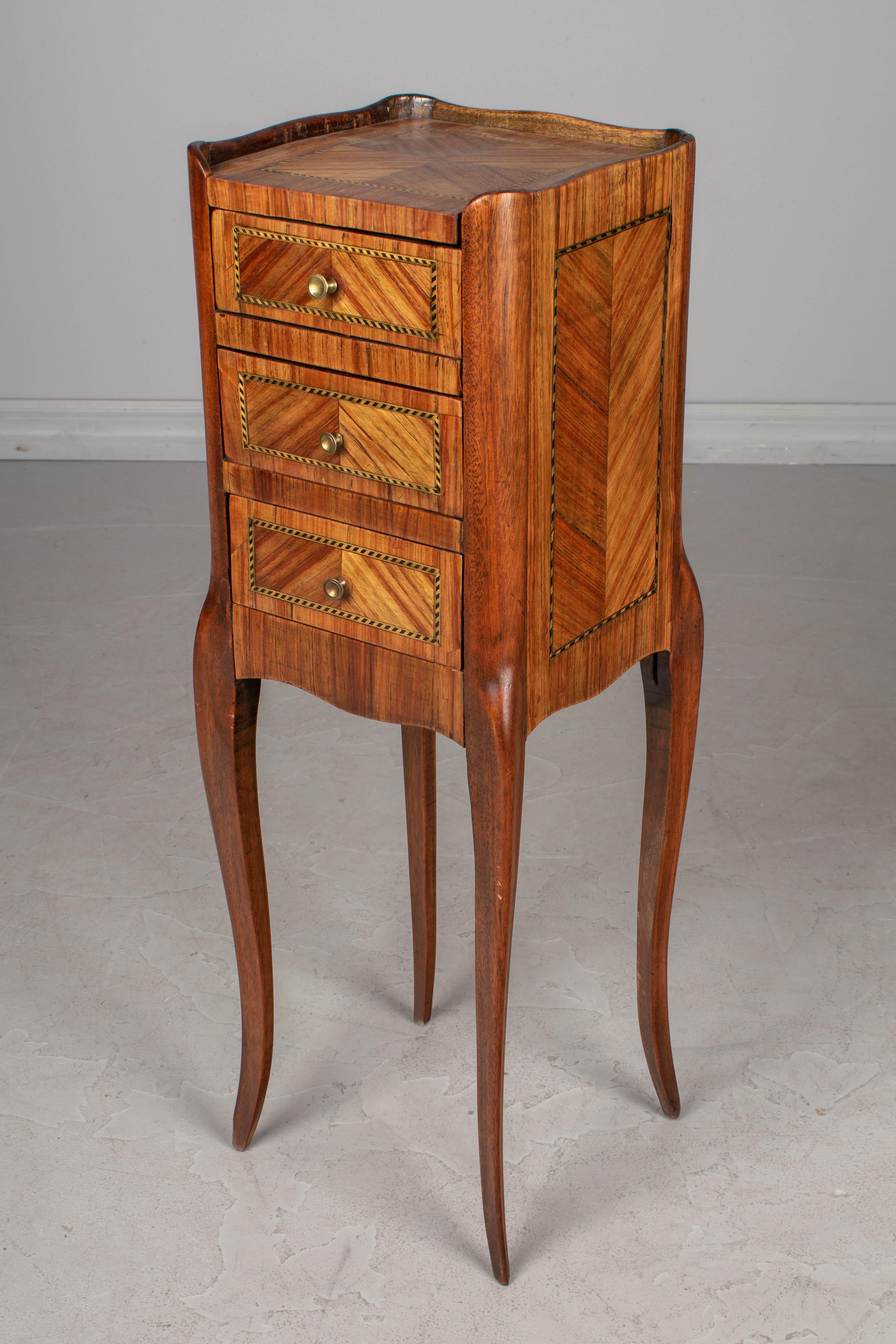 A small, elegant Louis XV style French marquetry side table, or night stand with veneers of walnut and mahogany inlaid in a chevron pattern. Three dovetailed drawers with brass pulls. Tall, slim proportions with curved corners and slender legs.