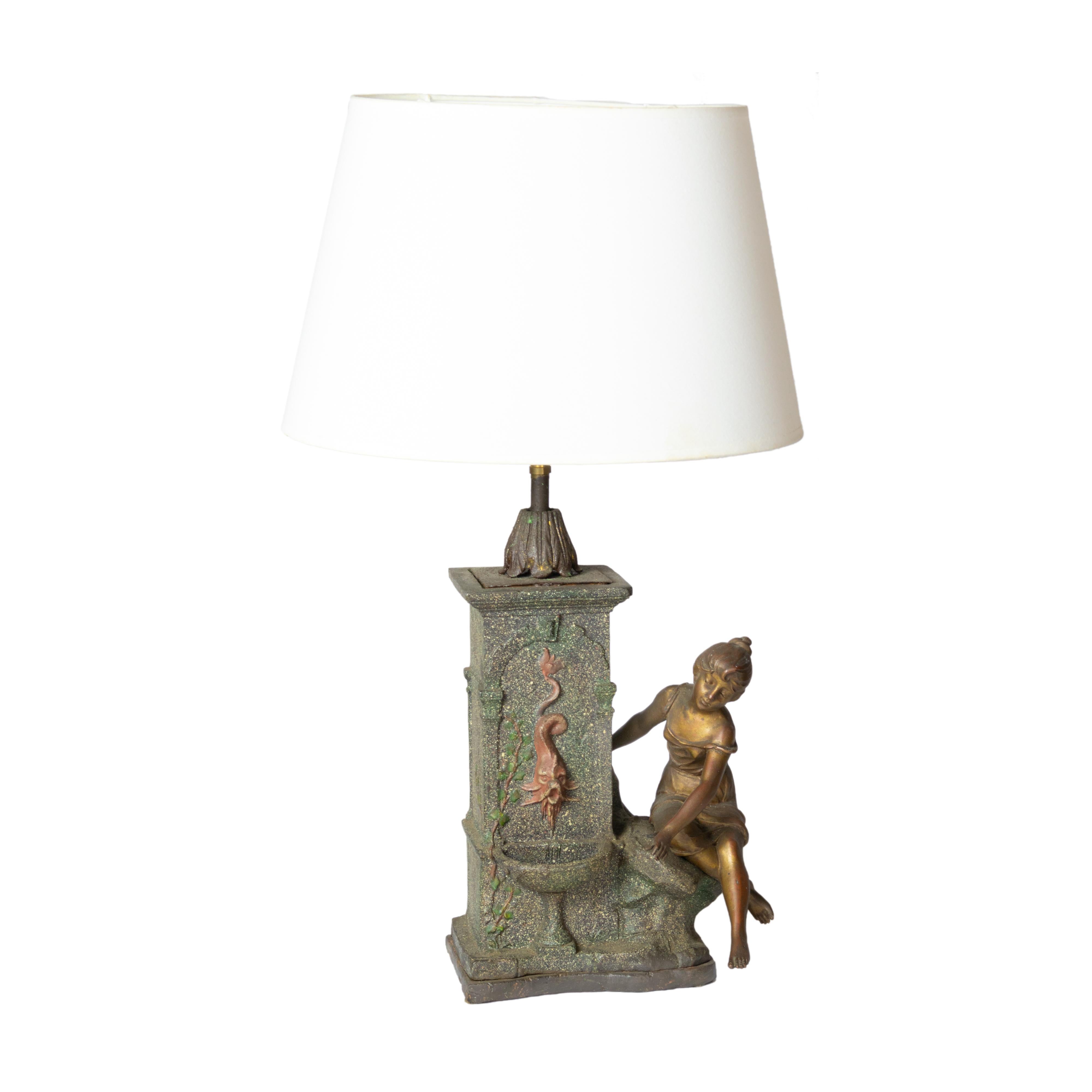 Une imposante lampe de table, ornée d'un vase décoratif, de bronze, d'éther et d'étain patiné, est placée en présence d'une jeune femme assise au bord d'une fontaine. La fontaine, ornée d'un dauphin, est dotée d'un plateau intérieur amovible en