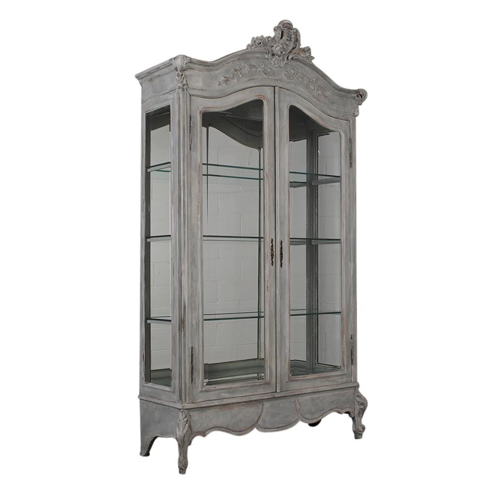 Two-door Louis XV-Style vitrine with beautiful carved details painted in a pale grey color with a distressed finish. The vitrine comes with beveled glass doors, three interior glass shelves, and a mirrored base and back. Working key and lock