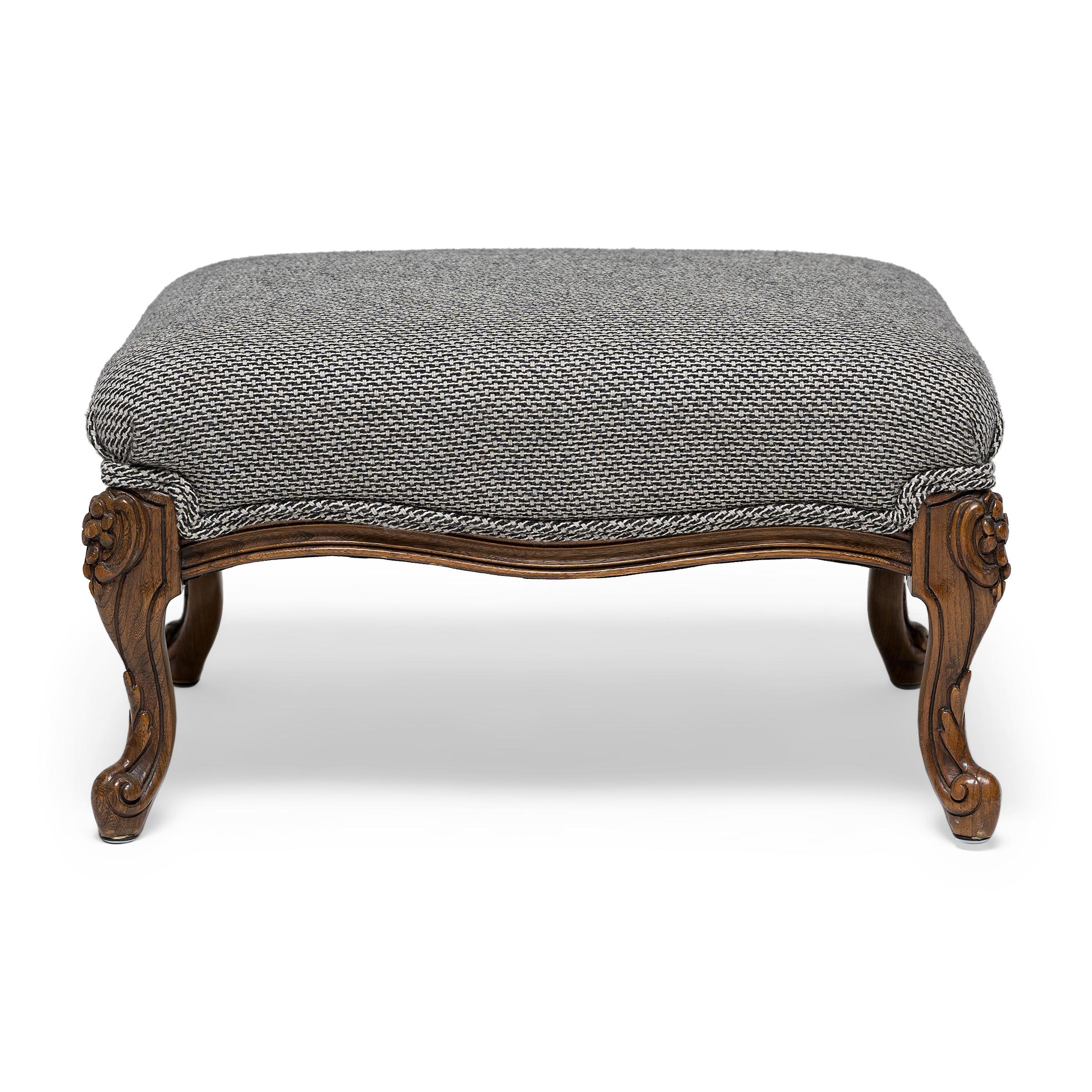 This rectangular footstool was crafted in 19th century France in the Louis XV style with an upholstered seat and an elegant carved fruitwood base. The stool rests on four short cabriole legs ending in dainty whorl feet, embellished with simple