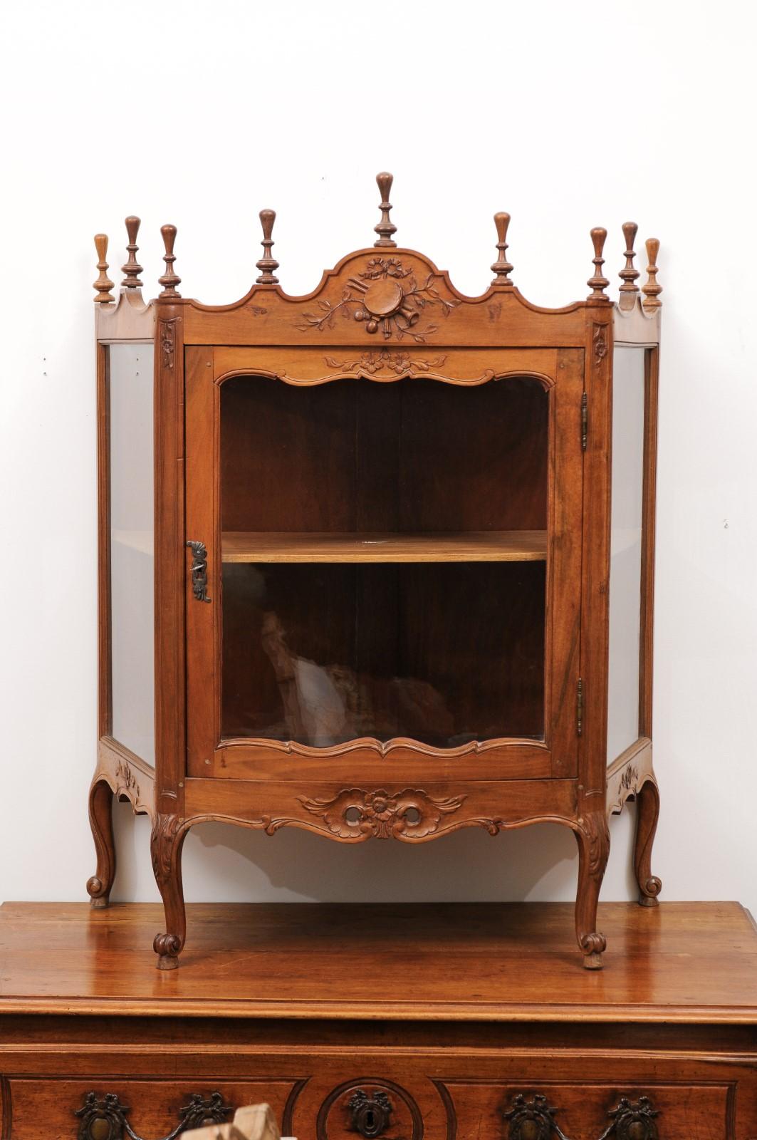 A French Louis XV style walnut display cabinet from the 19th century, with glass door, carved motifs and petite finials. Created in France during the 19th century, this Louis XV style petite vitrine captures our attention with its rhythmic