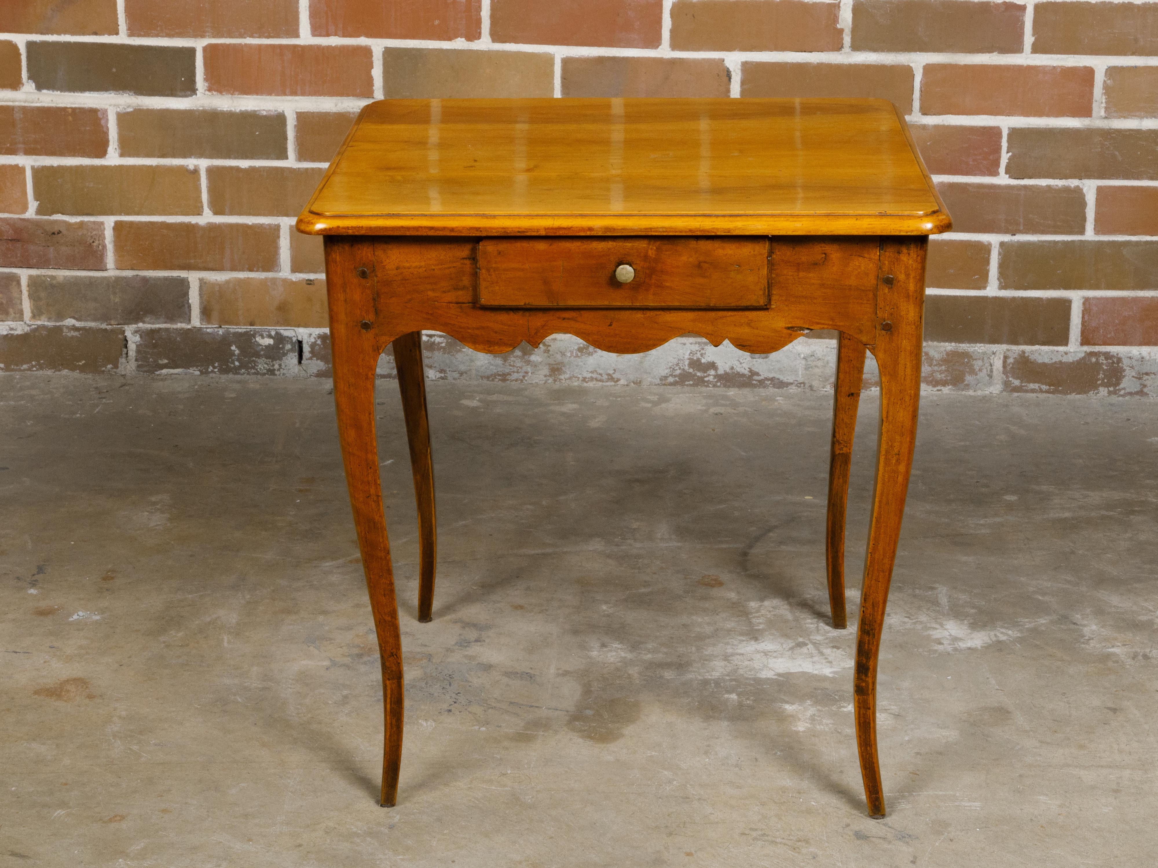 A French Louis XV style walnut side table from the early 19th century with single drawer and scalloped apron on all sides. This elegant French Louis XV style walnut side table, hailing from the early 19th century, exudes grace and sophistication.