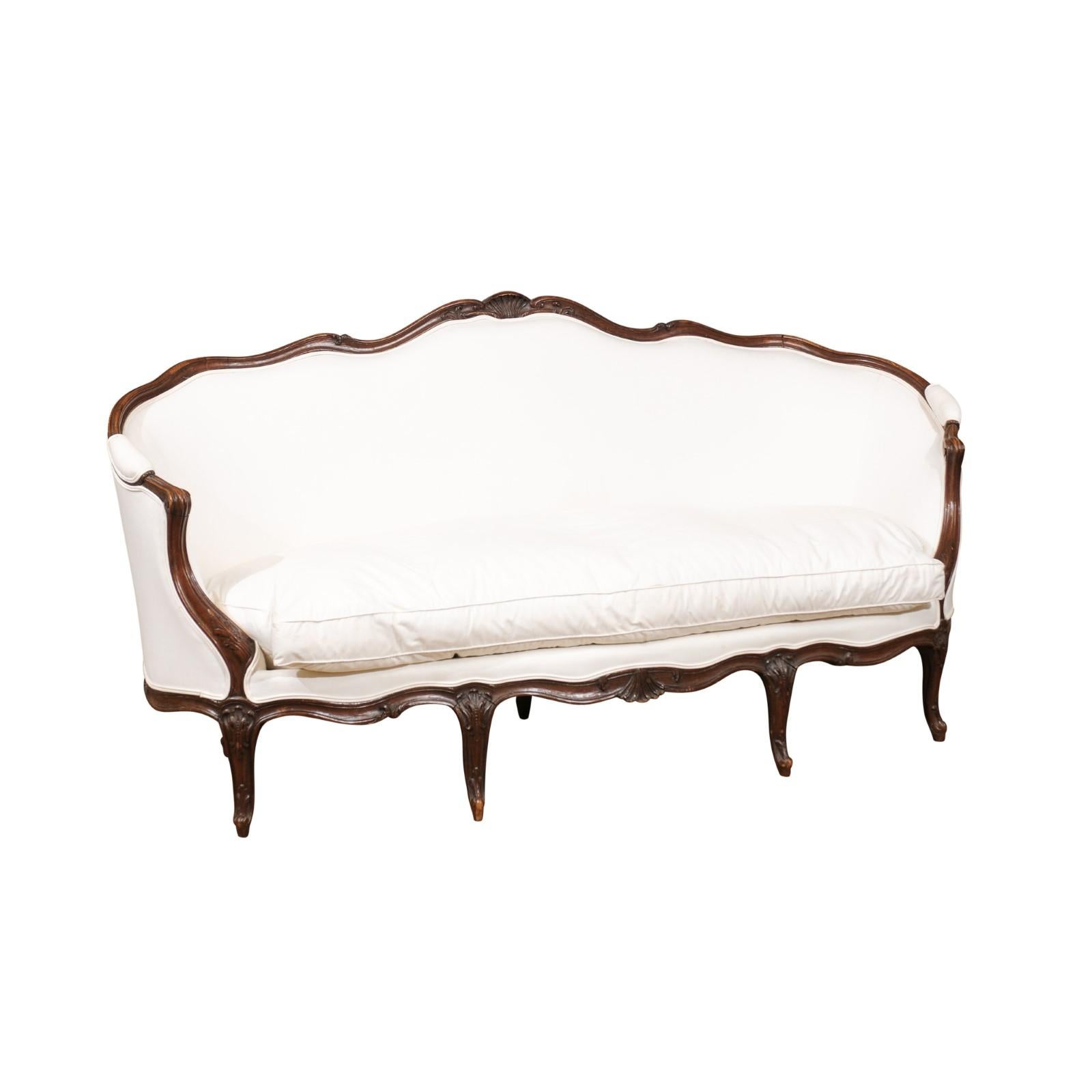 A French, Louis XV style walnut canapé from the mid-19th century, with wraparound back, scrolled arms, cabriole legs and new beige upholstery. This French canapé features a delicate wraparound back, adorned with a shell-carved crest as well as tall,