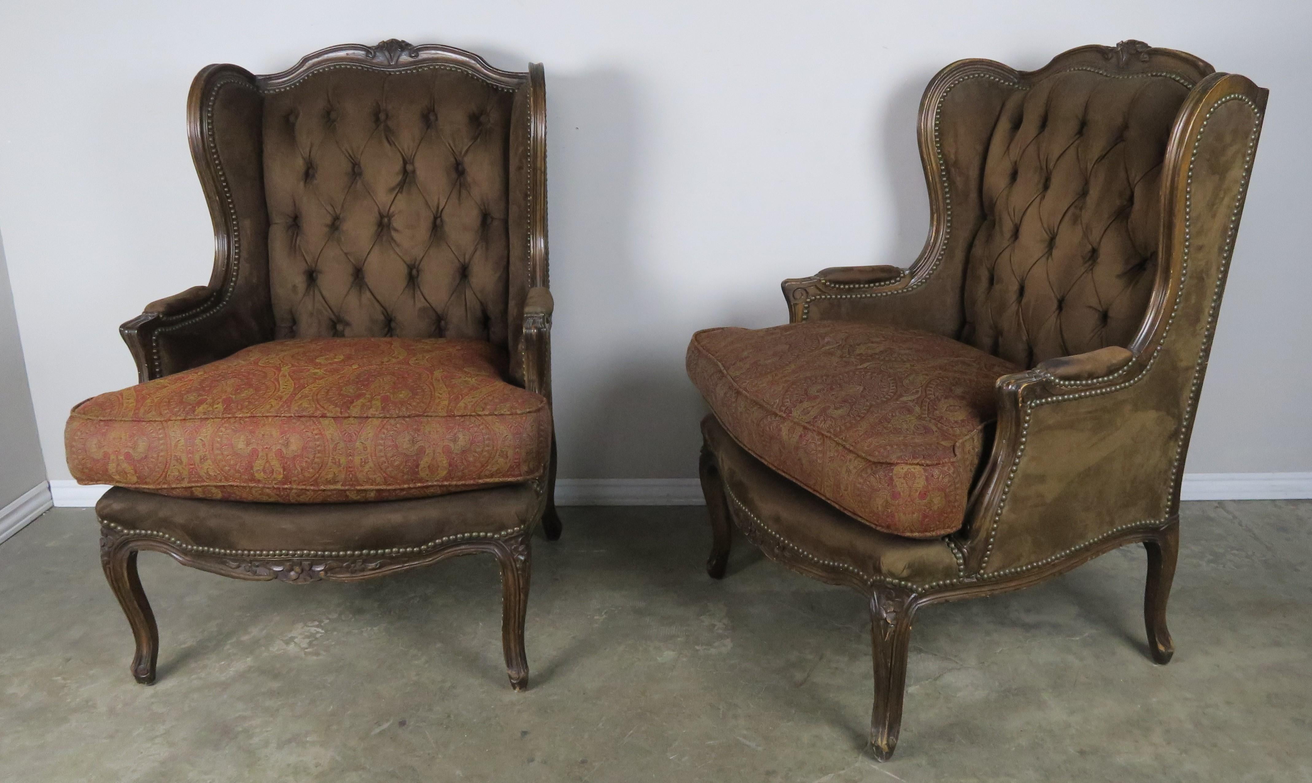 Pair of French Louis XV style carved walnut wingback armchairs upholstered in tufted ultra suede with antique brass colored nailhead trim detail. The armchairs stand on four cabriole legs. Loose contrasting cushions upholstered in a wool paisley