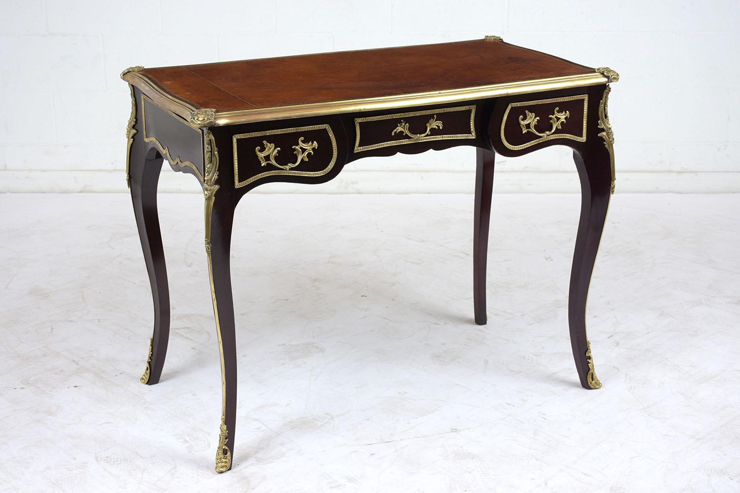 This 1900s Louis XV-style desk is made of mahogany wood stained a dark mahogany color with a lacquered finish. The top of the desk features embossed leather with original distressed finish, along with a detailed brass molding around the top. There