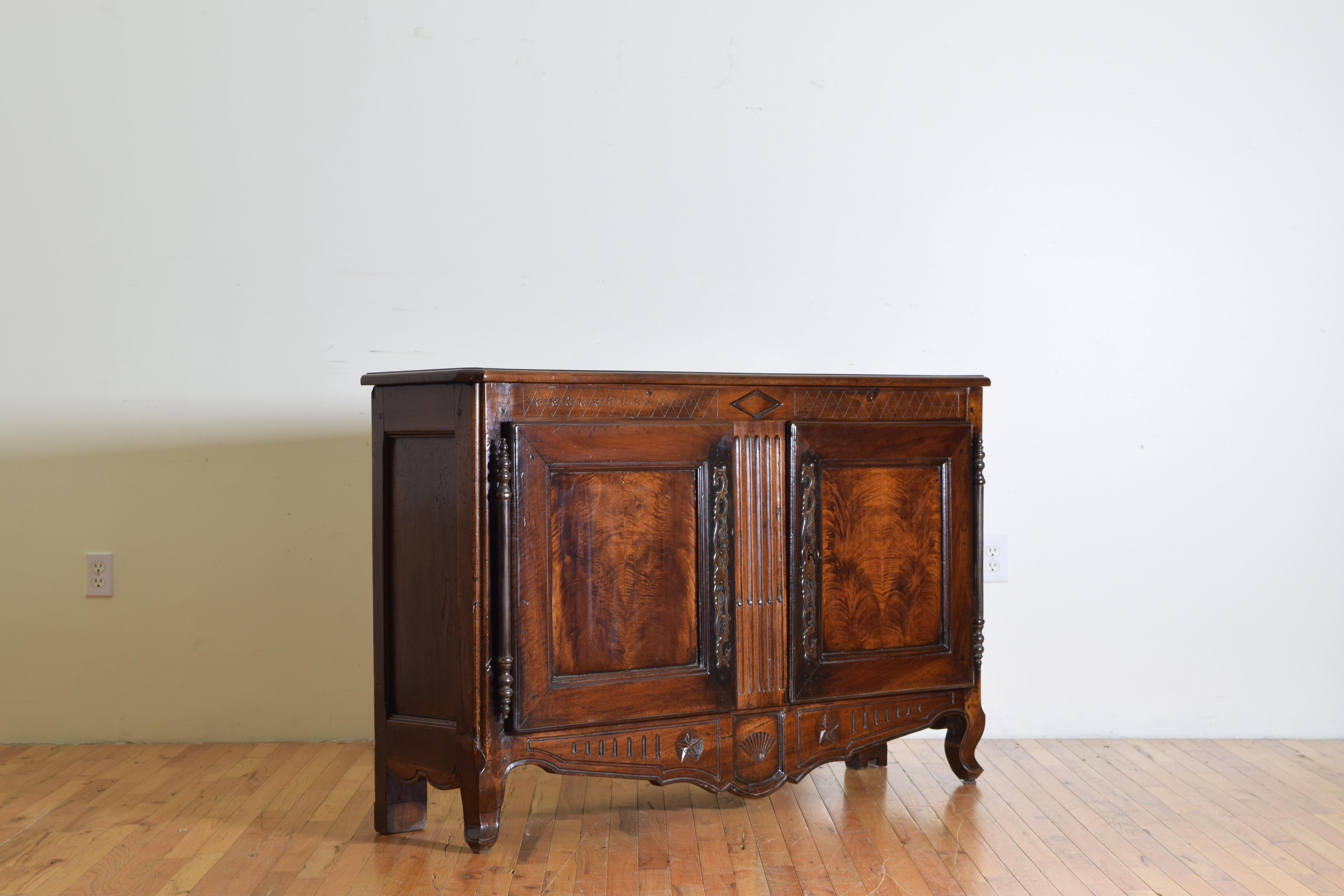 Having a rectangular top with canted front corners above a conforming case with two doors mounted on large steel hinges, upper diagonal carvings with a centered horizontal lozenge, matched veneer paneled doors, the center section with vertical
