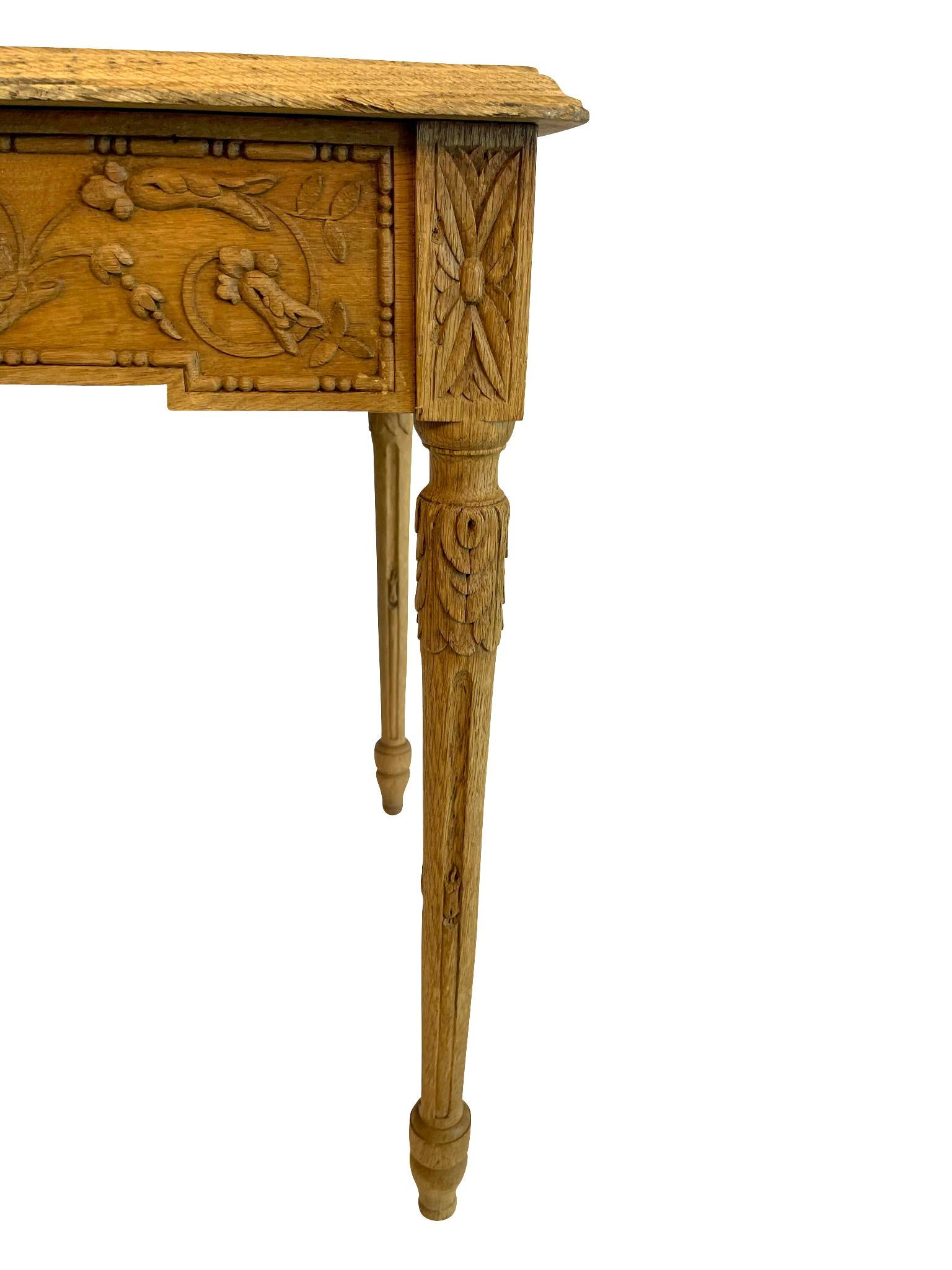 A lovely French Louis XVI bleached oak side table on tapered fluted legs with a carved apron of neoclassical design—Laurel wreaths in a foliate pattern with garlands of hanging ribbons and bows. The corners have the classic carved rosette design,