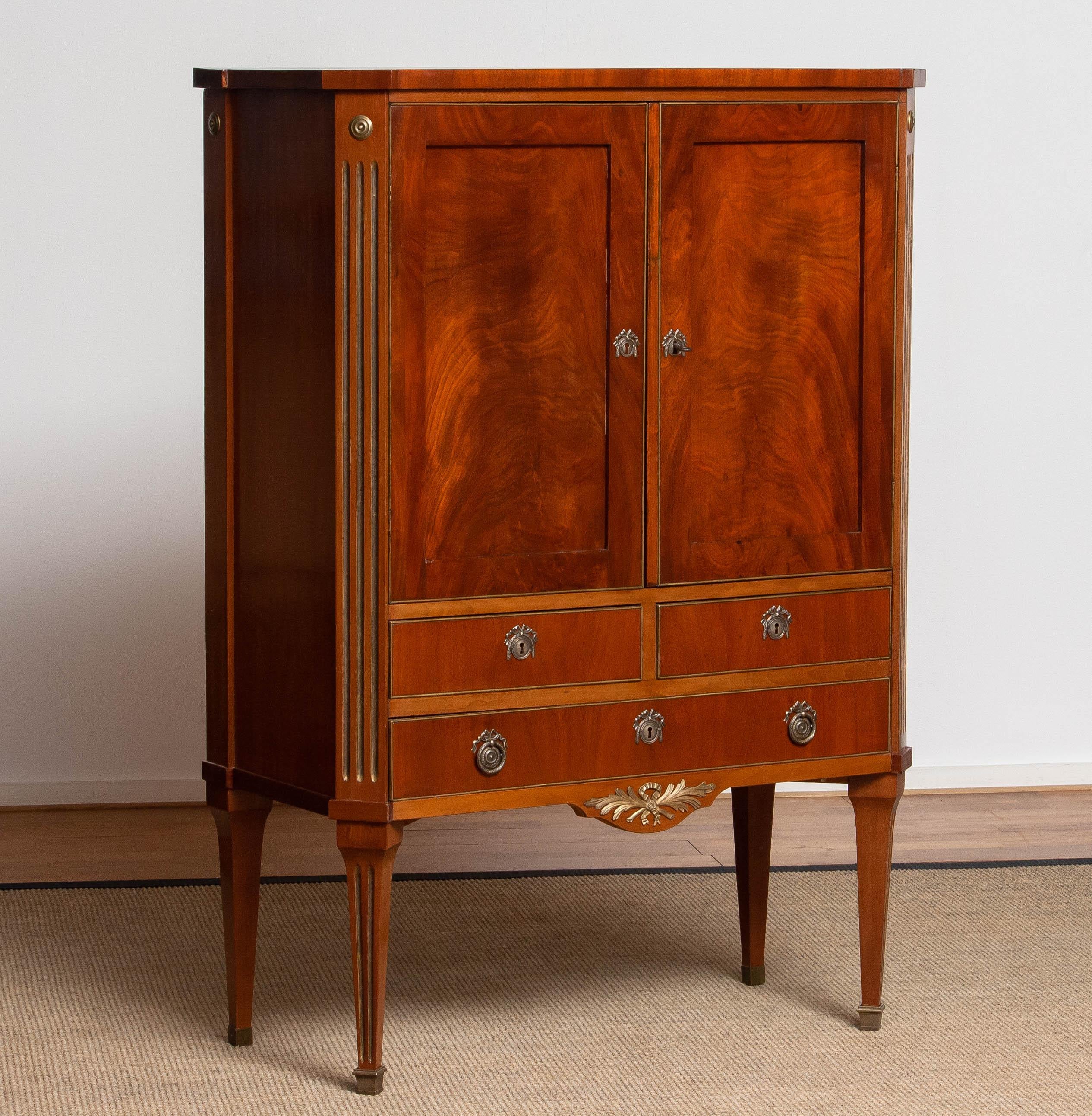 Elegant Louis XVI style cabinet from France, crafted in mahogany veneer with refined brass inlays. The cabinet features two paneled doors and three drawers, all adorned with brass detailing and original decorative brass hardware. Fluted side columns