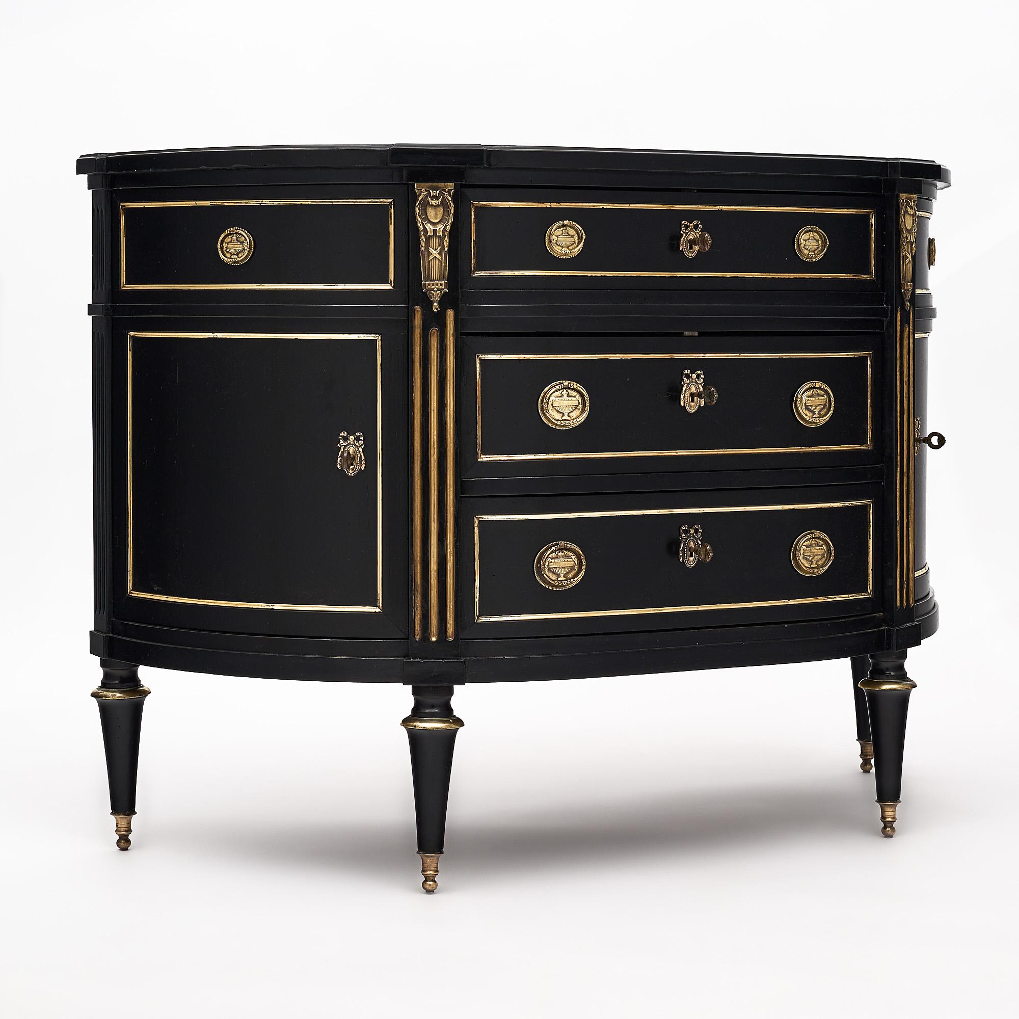 Demilune chest of drawers, French, in the Louis XVI style mad of solid ebonized cherrywood and finished with a lustrous Museum quality French polish. There are three dovetailed drawers, two doors, gilt brass trim throughout, and finely cast hardware.