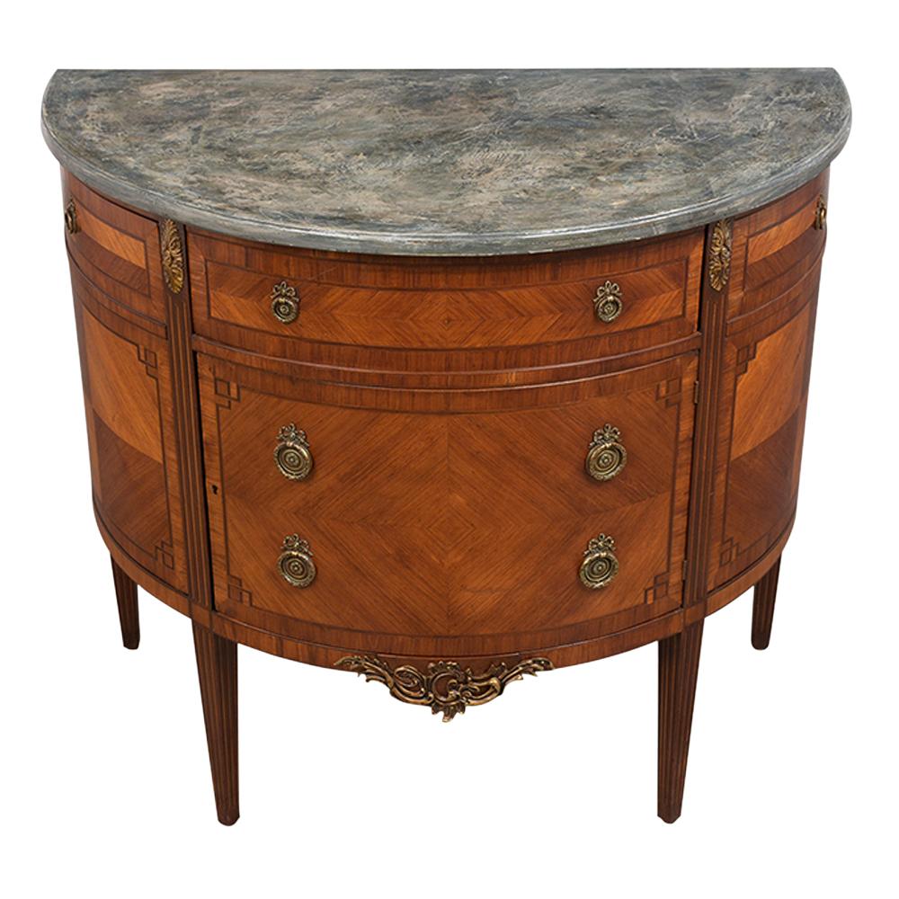 This Vintage French Louis XVI Style Demilune Commode is in excellent condition has been finished in a rich mahogany color and features a beautiful inlaid fruitwood veneer. The commode also features a faux marble top with a beveled edge, the bottom