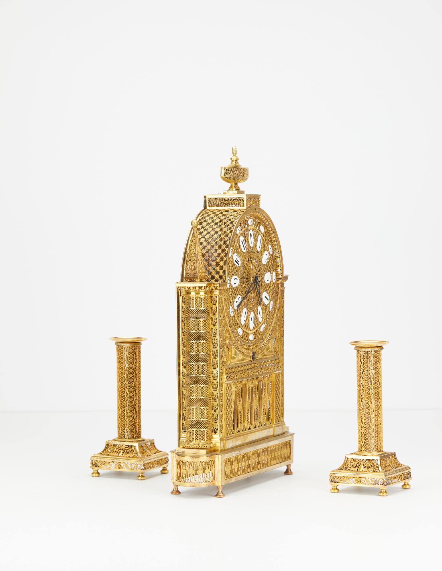 L'ensemble comprend une horloge squelette en bronze doré de style Arabesque, ainsi que les chandeliers d'origine en bronze doré assortis. L'horloge repose fièrement sur quatre pieds et une base filigranée. Le cadran Arabesque très détaillé comporte