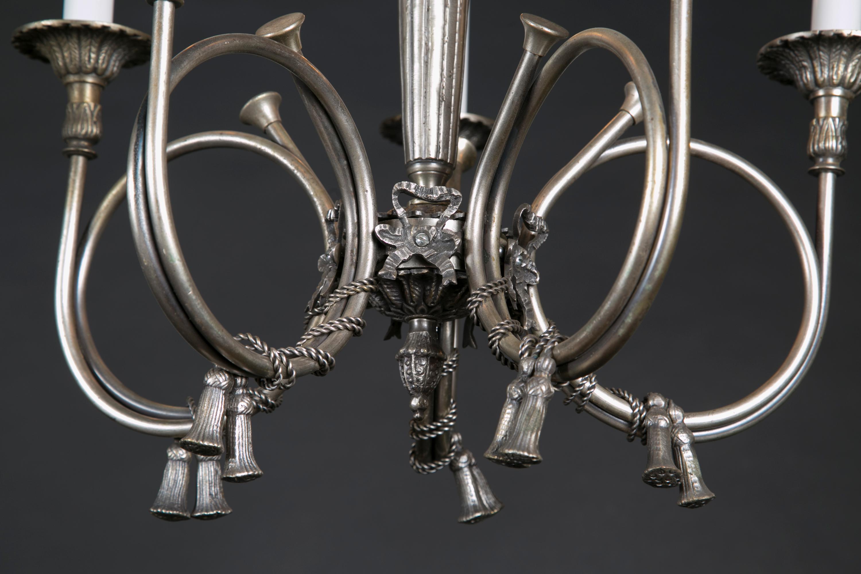 This French Belle Epoque chandelier is made of silvered brass, and features beautiful ‘French Horn’ arms draped with detailed rope and hung with tassels. The piece dates back to the early 20th century, and features acanthus leaves on top the fluted