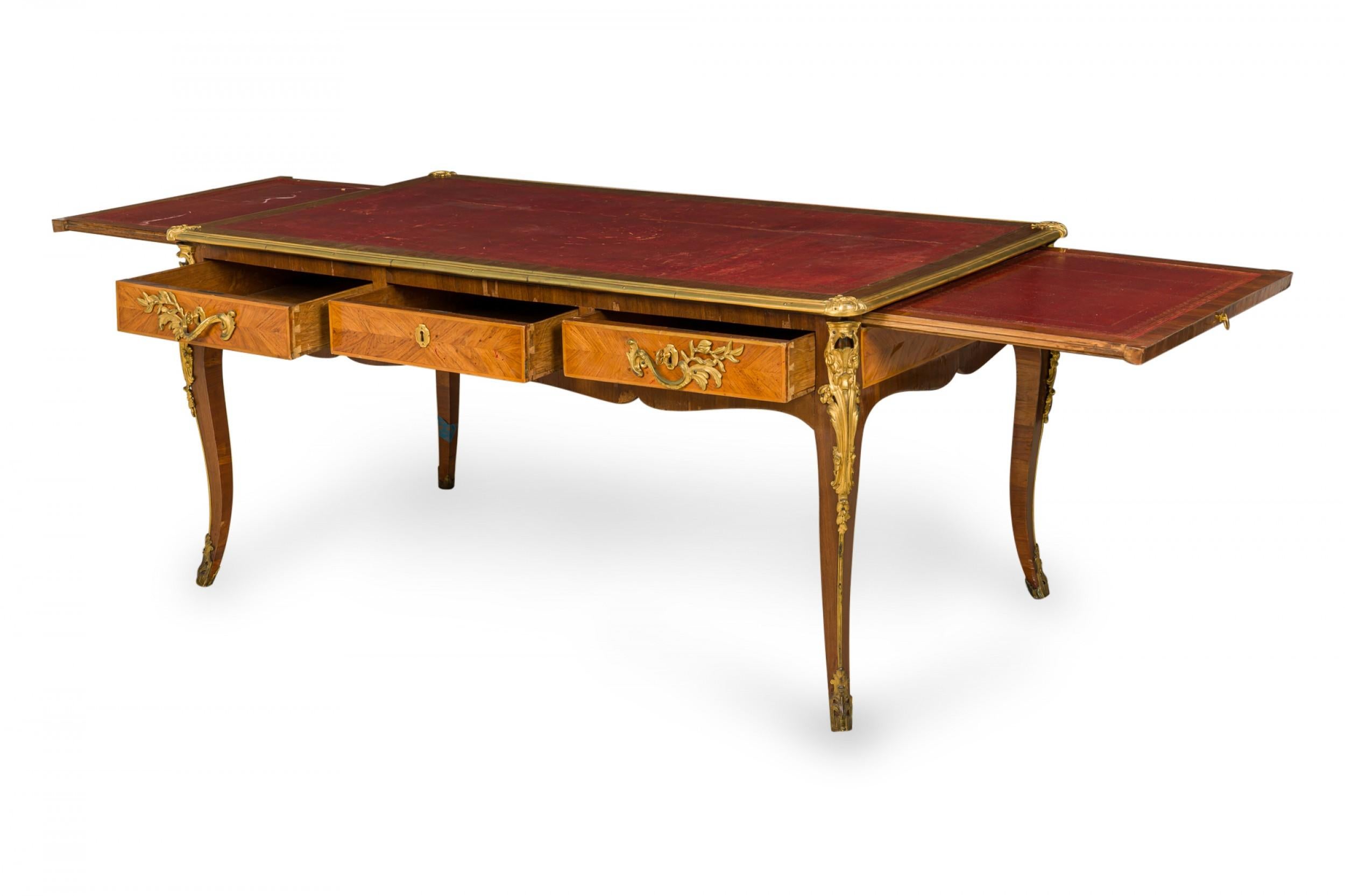 French Louis XVI style (19th century) writing desk with kingwood veneer, a rectangular red leather desktop with embossed gold border and brass trim, with two extendable writing surfaces over three drawers with two elaborate ormolu drawer pulls a
