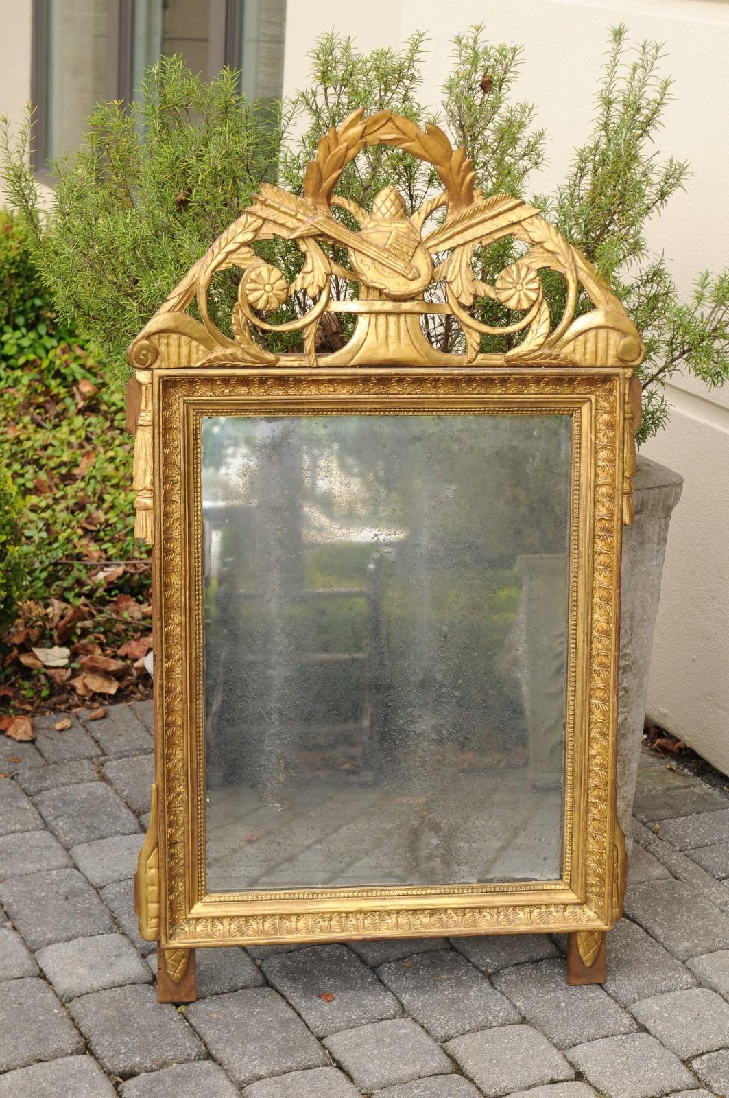Ce miroir en bois doré sculpté de style Louis XVI de la fin du XVIIIe siècle présente un exquis cimier ajouré et sculpté composé de divers motifs tels qu'un luth, un gland, des flèches, des fleurs, une couronne de laurier et des volutes symbolisant