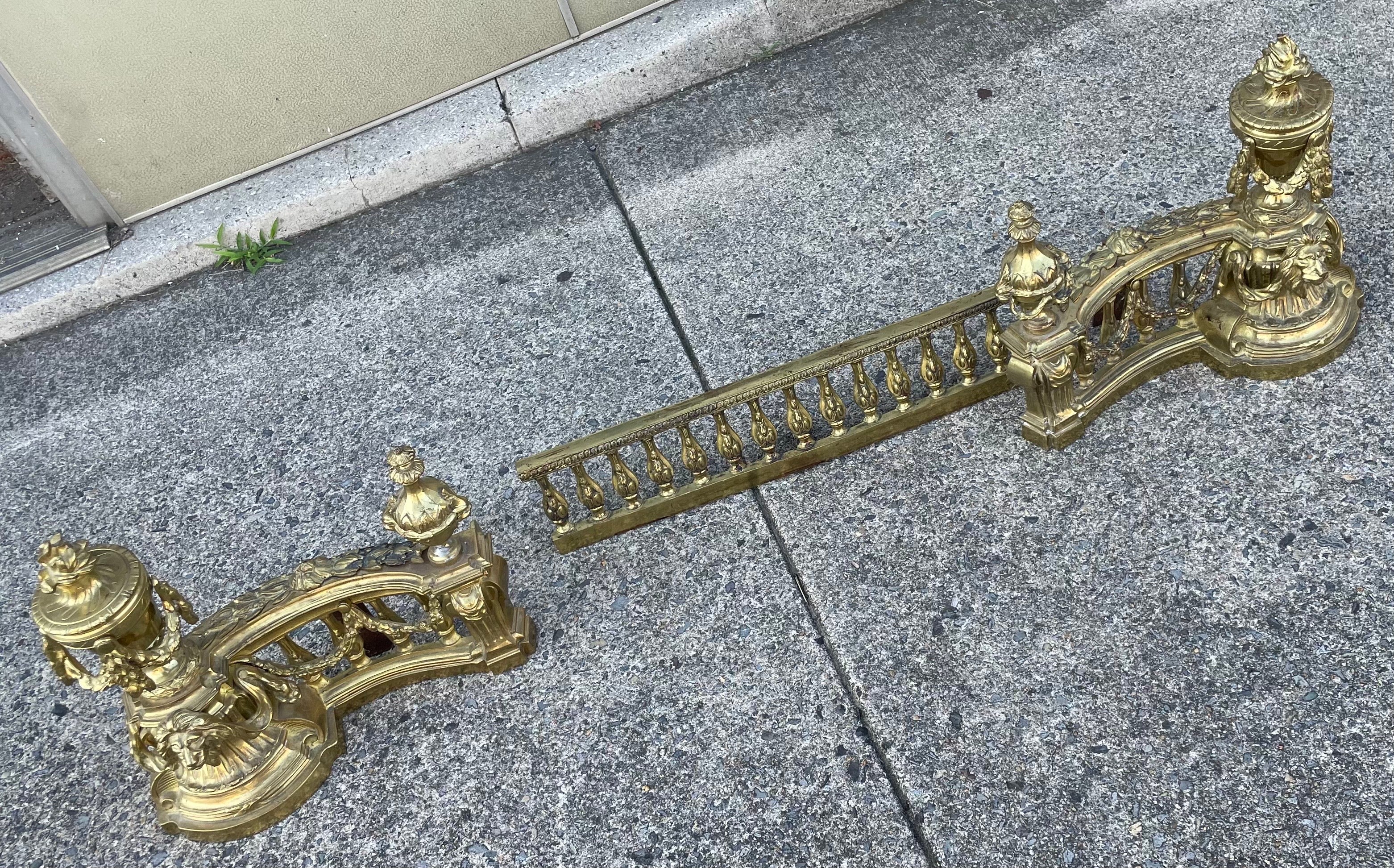 This is a fantastic set of Louis XVI style chenets with the connecting fender that have wonderful neoclassical style elements including swag and acanthus leaf decorations, a protecting lion's head and urns with a flame on top, the gold gilt brass