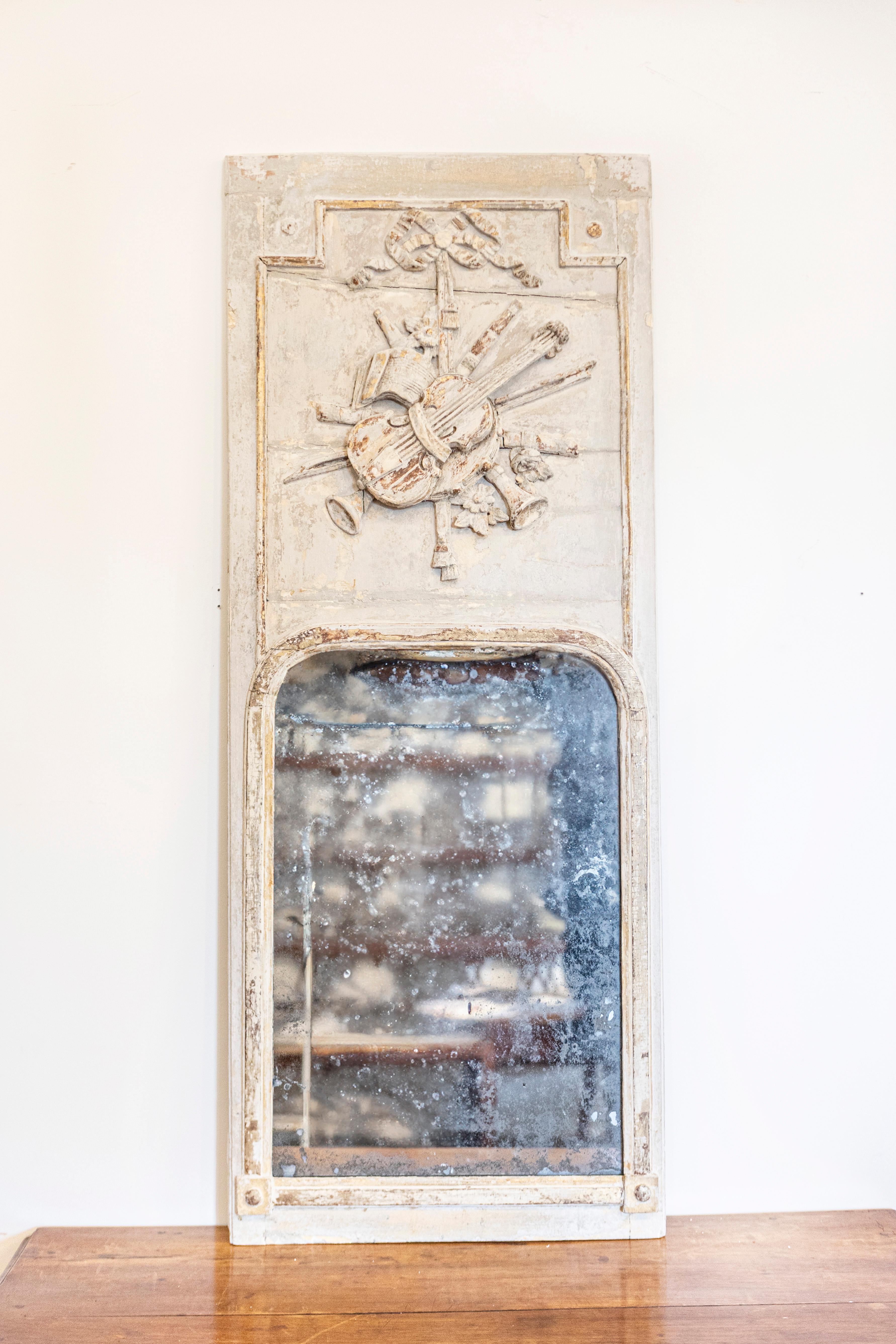 Espejo trumeau francés de época Luis XVI de hacia 1790 con alegoría de las Artes Liberales tallada, acabado pintado en blanco hueso antiguo y detalles dorados. Este cautivador espejo trumeau francés de época Luis XVI, de hacia 1790, es un emblema de