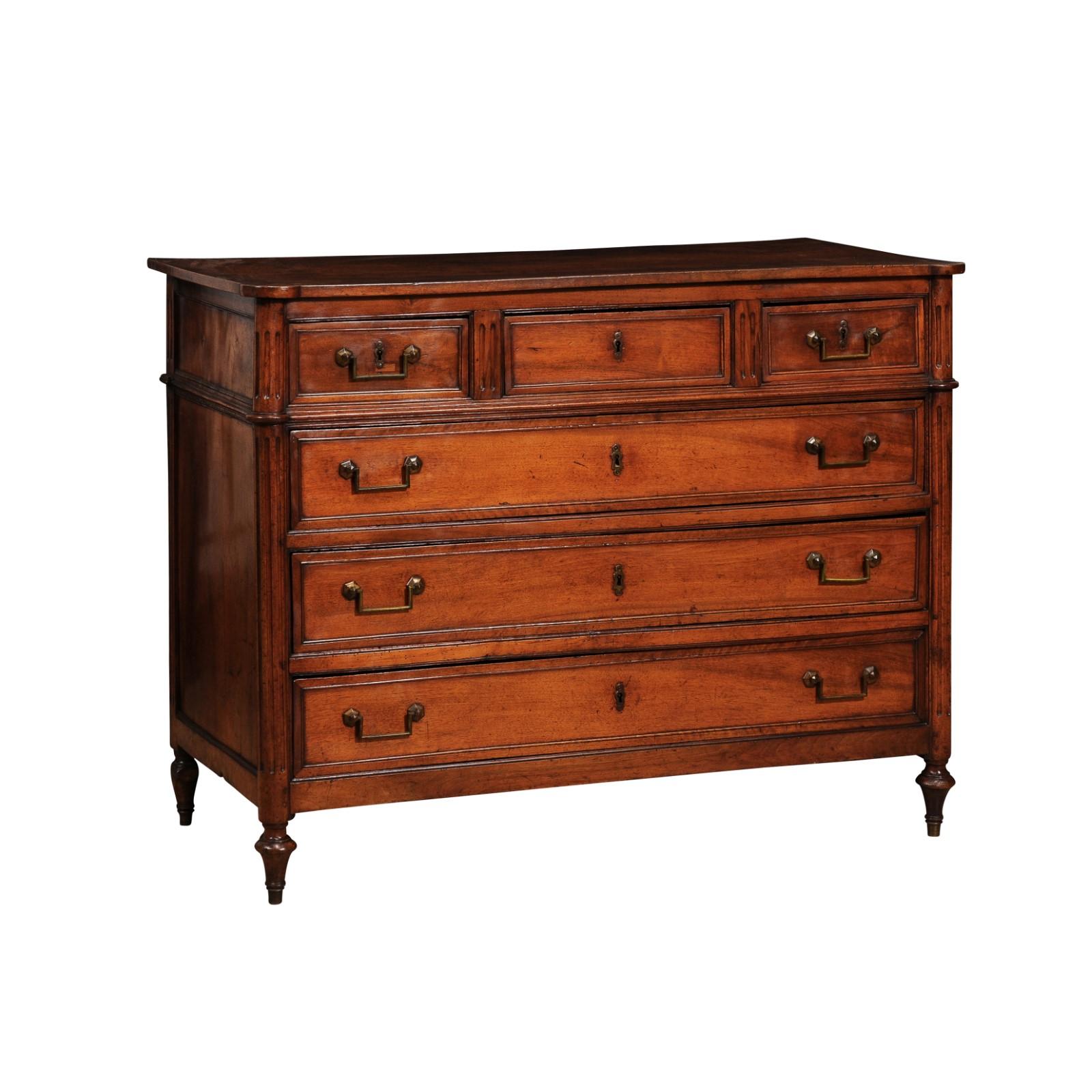 A French Louis XVI period walnut commode from circa 1790 with six drawers, rounded and fluted side posts, and turned elongated toupie legs. This French Louis XVI period walnut commode, dating back to circa 1790, is an exquisite representation of