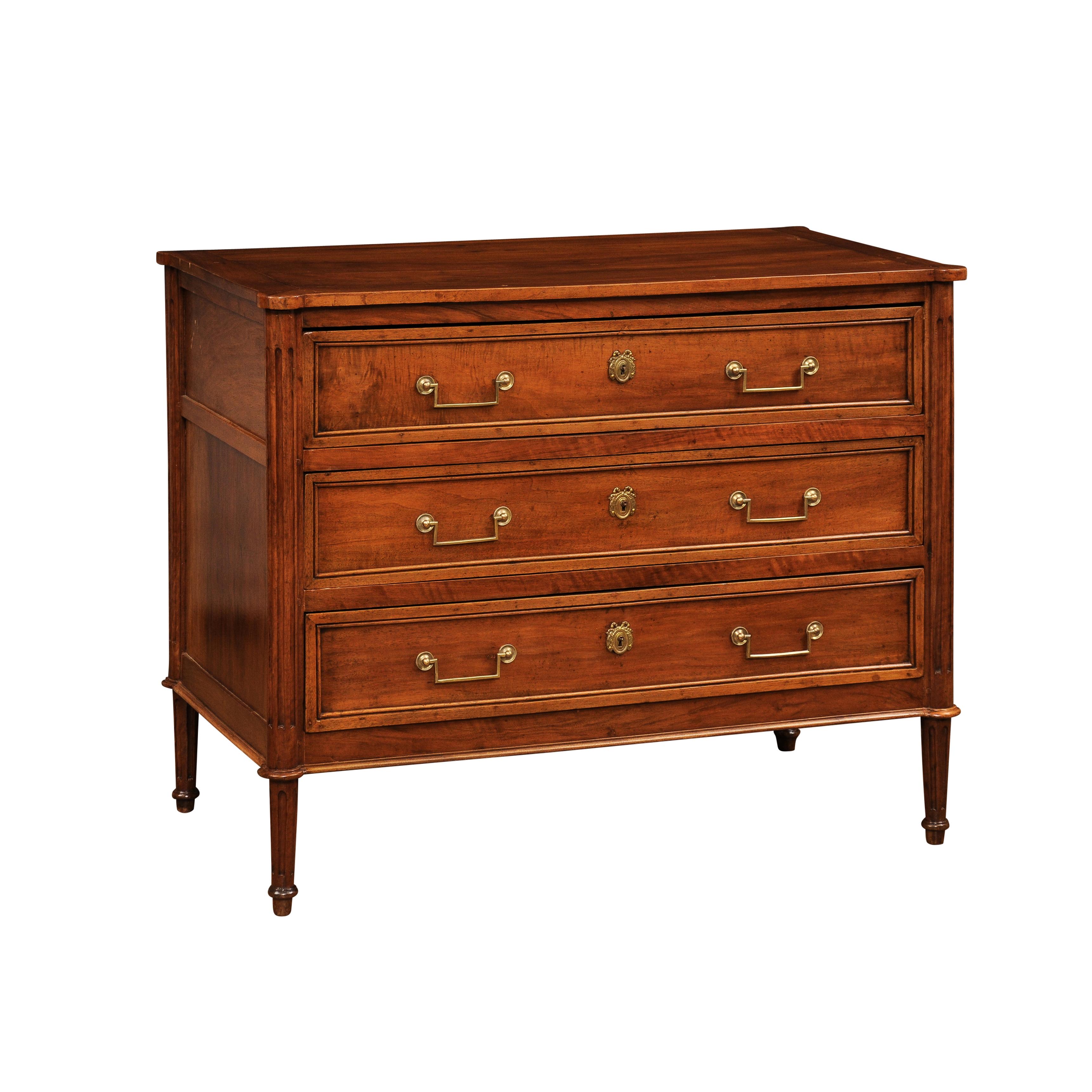 A French Louis XVI period walnut commode circa 1790 with three drawers, linear brass hardware, cylindrical tapered legs and rounded fluted side posts. Step into the elegance of the French Louis XVI period with this exquisite walnut commode from the